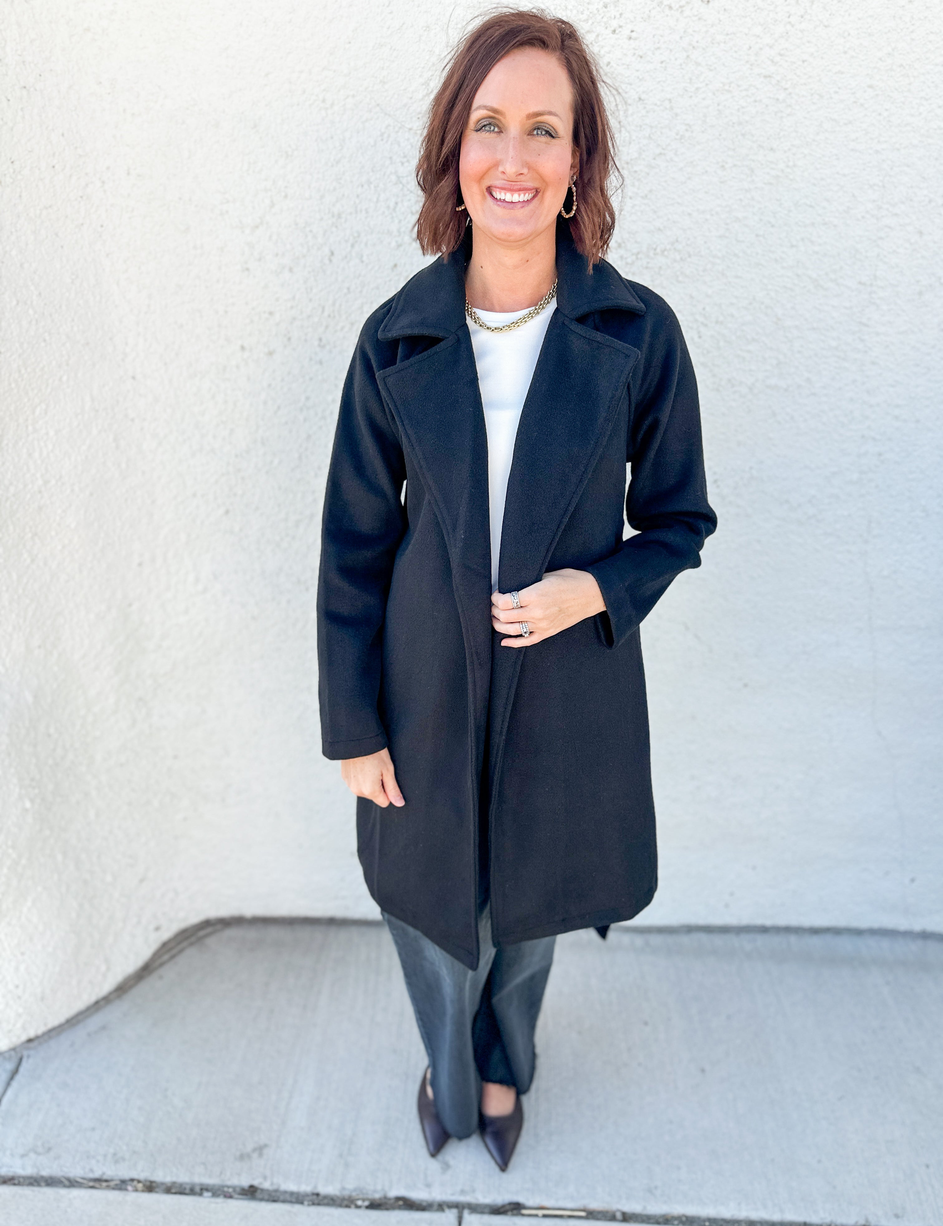 A woman stands outdoors against a light textured wall wearing a long black coat over a white top and gray pants.