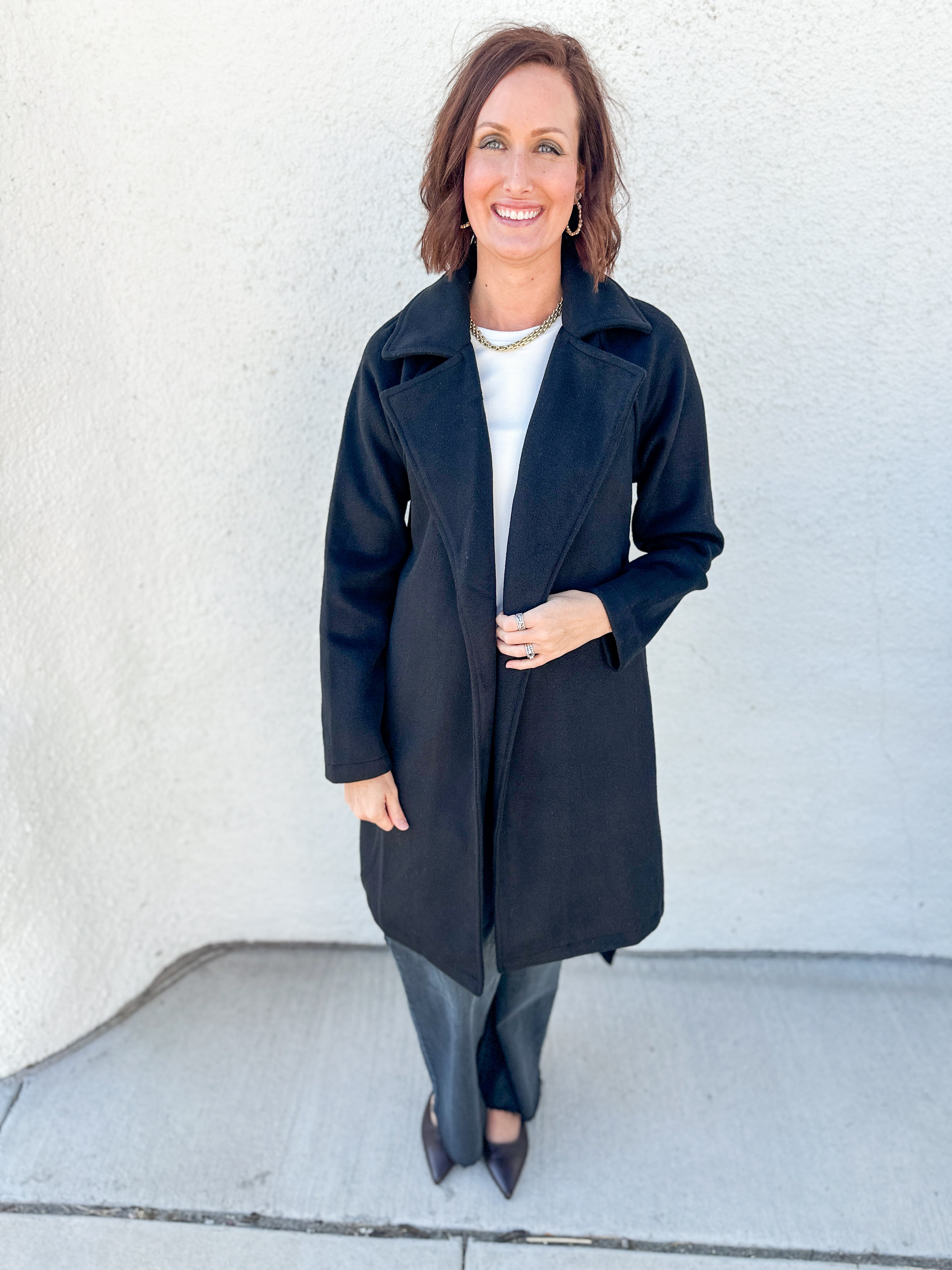 A woman stands outdoors against a light textured wall wearing a long black coat over a white top and gray pants.