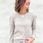 A woman wearing a light taupe long-sleeve lightly ribbed crewneck sweatshirt with ruched shoulder detail and matching taupe sweatpants, standing against a textured white wall.