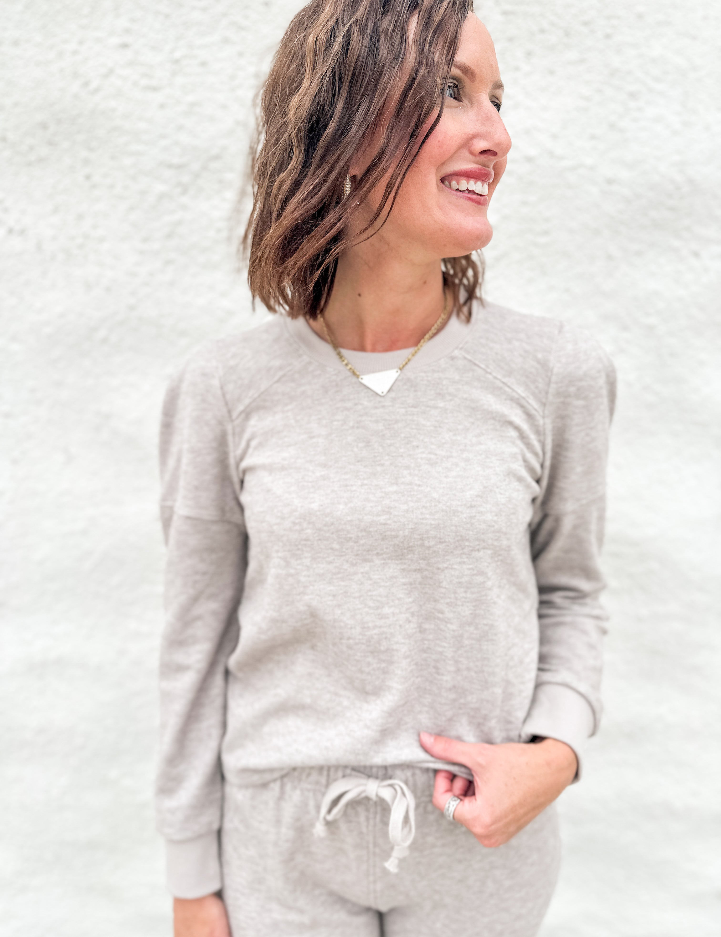 A woman wearing a light taupe long-sleeve lightly ribbed crewneck sweatshirt with ruched shoulder detail and matching taupe sweatpants, standing against a textured white wall.