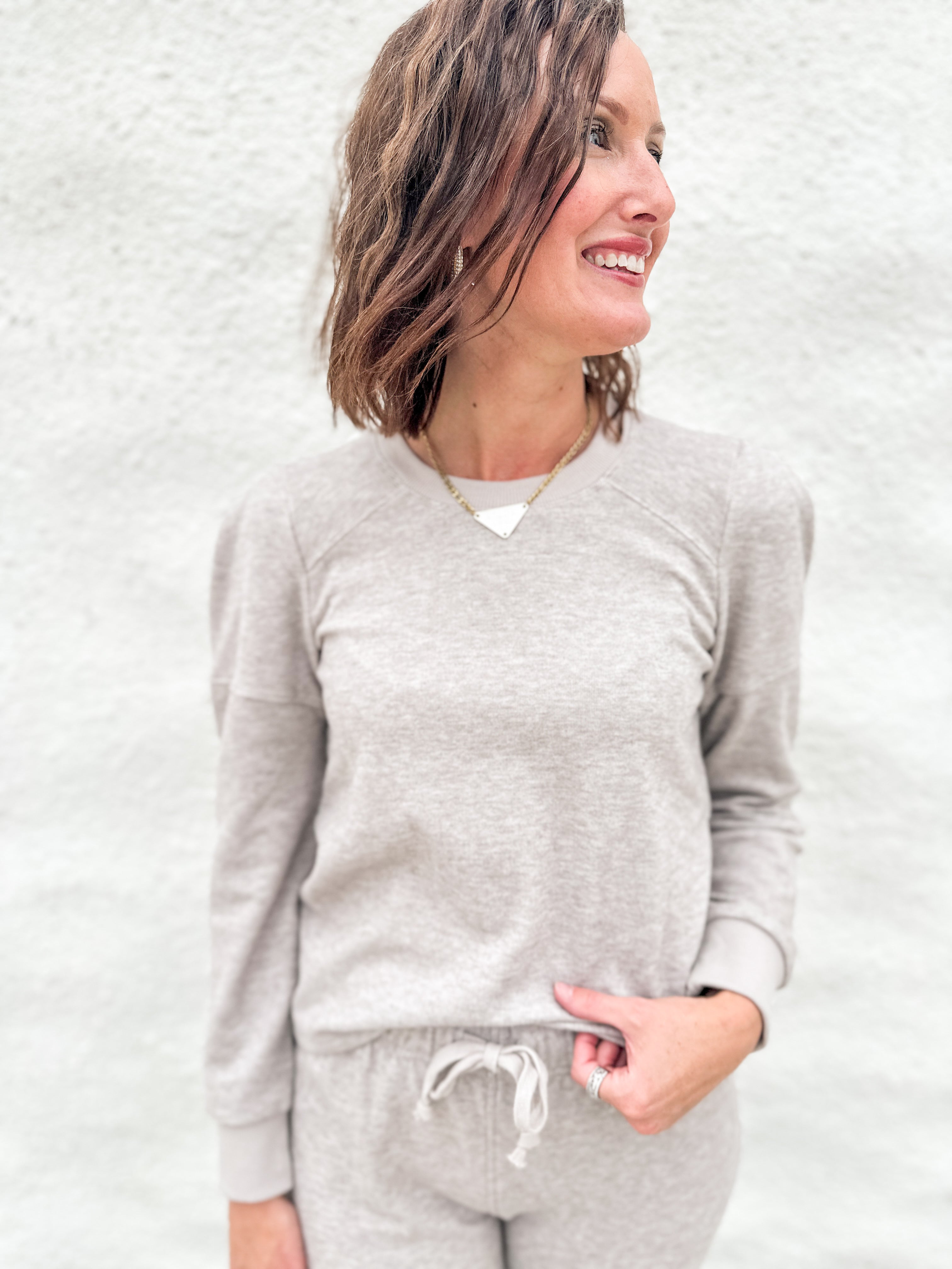 A woman wearing a light taupe long-sleeve lightly ribbed crewneck sweatshirt with ruched shoulder detail and matching taupe sweatpants, standing against a textured white wall.