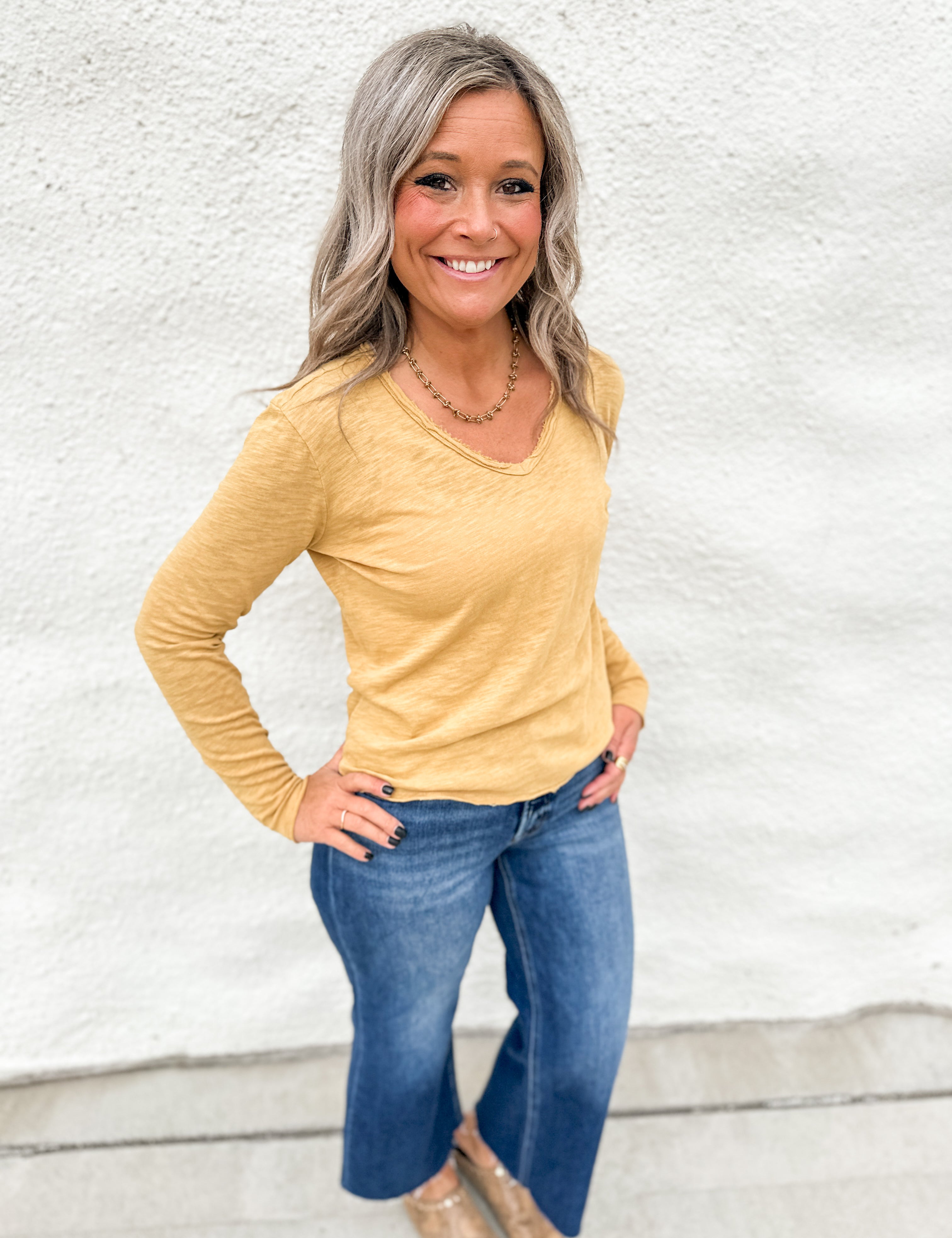A woman wearing a long sleeve top with rolled hems wearing blue jeans standing in front of a white wall.
