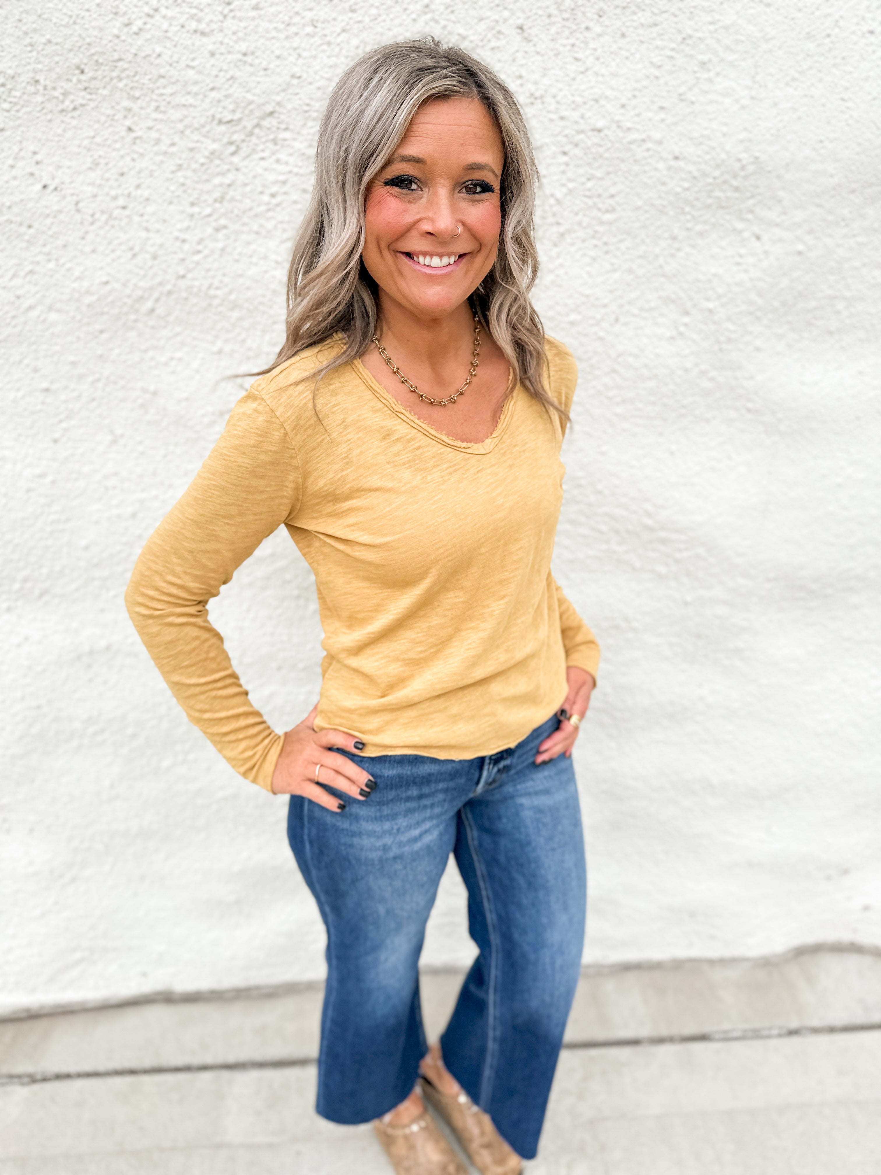 A woman wearing a long sleeve top with rolled hems wearing blue jeans standing in front of a white wall.