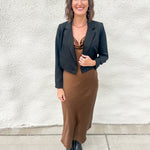 A woman stands outdoors against a textured white wall wearing a black cropped blazer over a brown satin-like top and a brown midi skirt, paired with black pointed shoes.