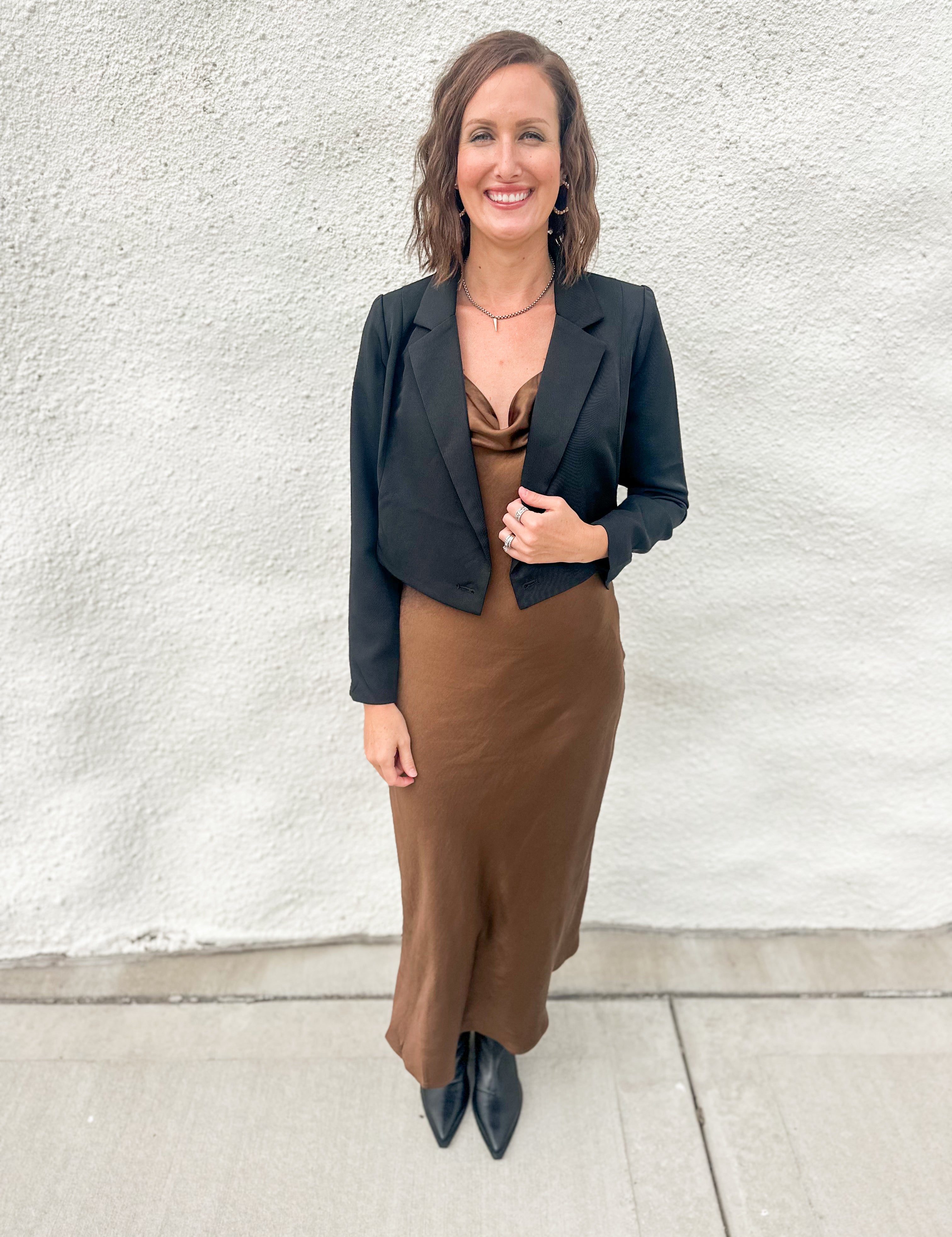 A woman stands outdoors against a textured white wall wearing a black cropped blazer over a brown satin-like top and a brown midi skirt, paired with black pointed shoes.