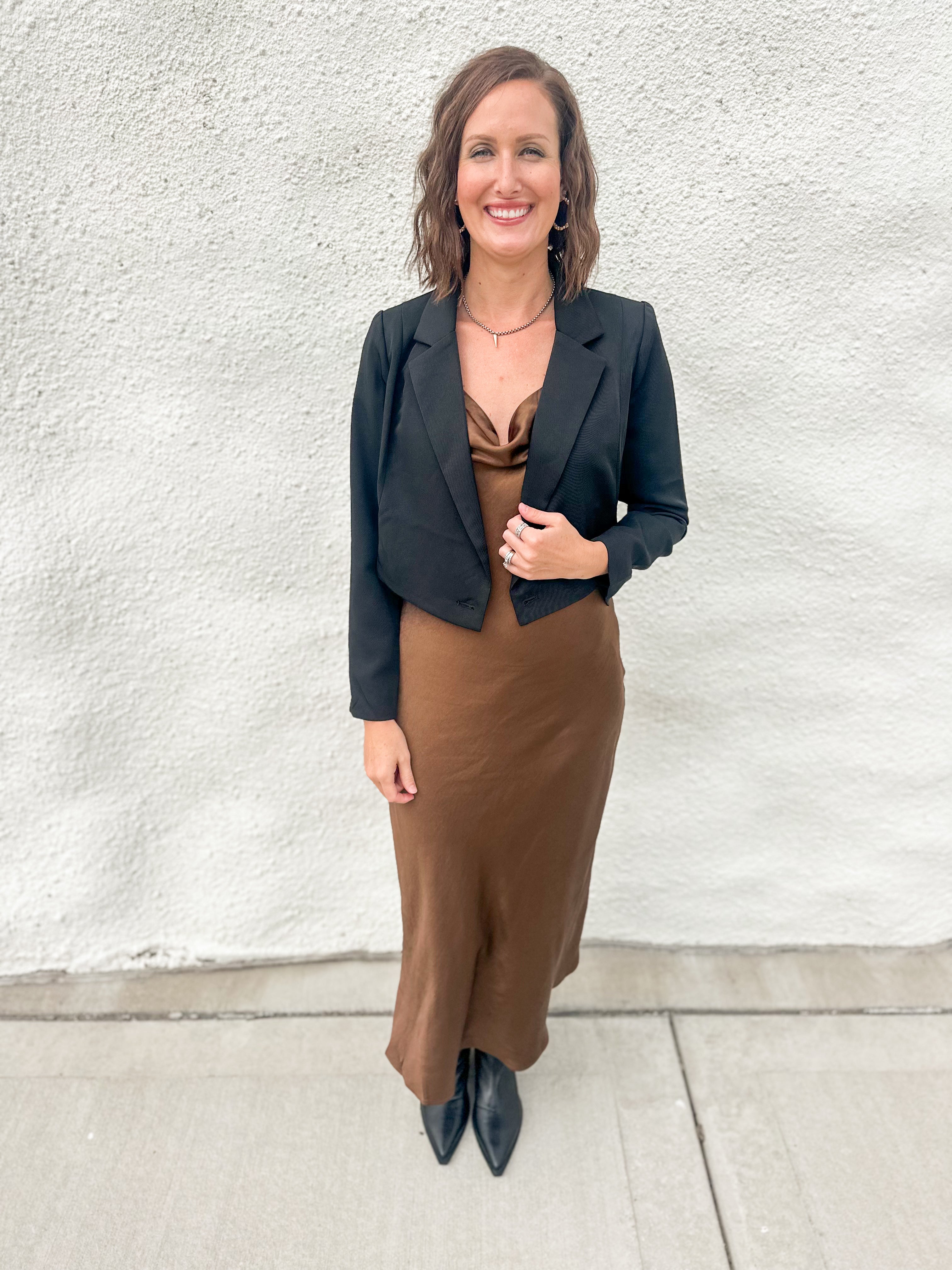 A woman stands outdoors against a textured white wall wearing a black cropped blazer over a brown satin-like top and a brown midi skirt, paired with black pointed shoes.