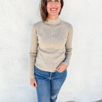 A woman smiling and wearing a beige mock neck knit sweater and blue jeans, standing against a white textured wall.