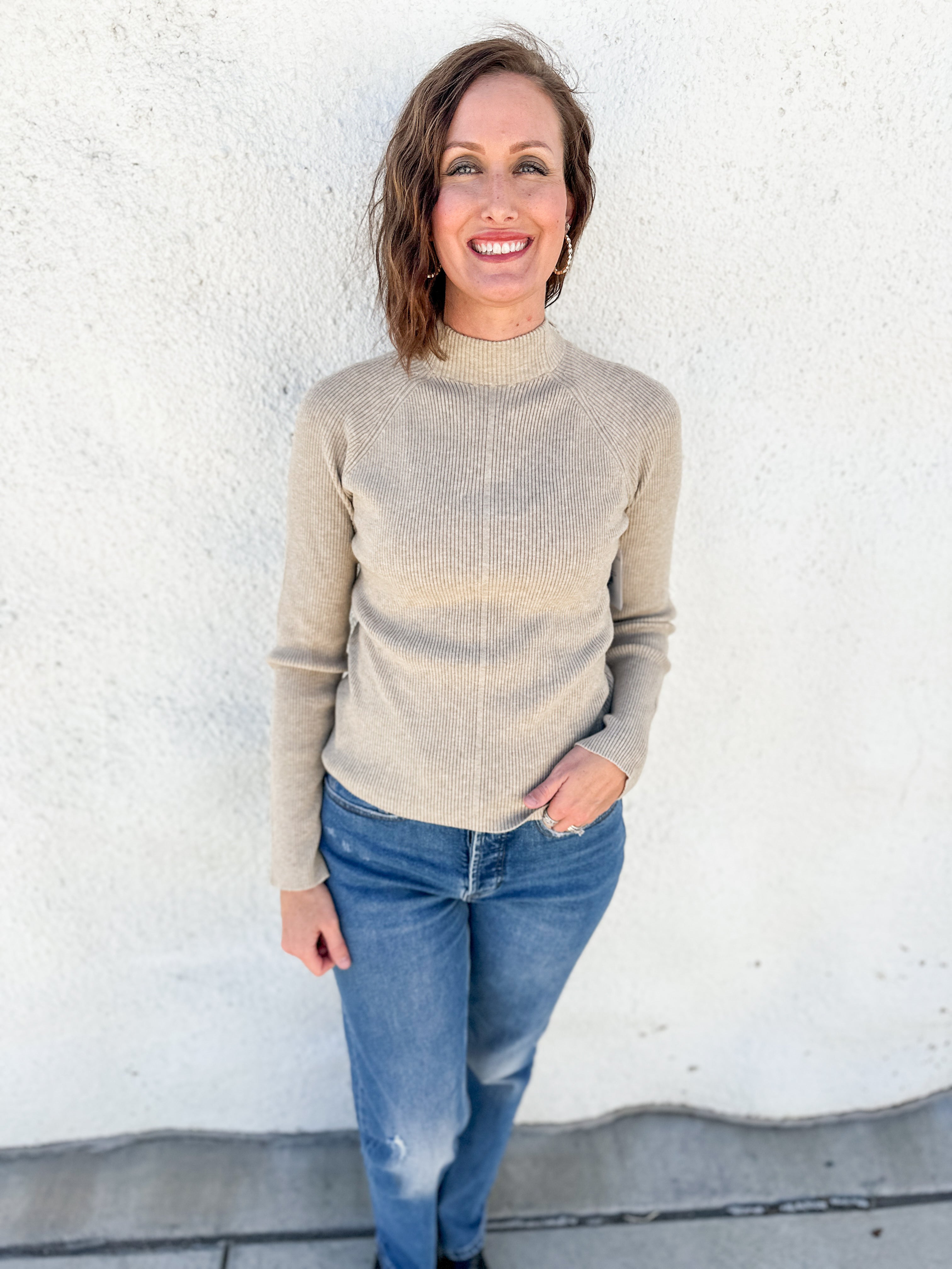 A woman smiling and wearing a beige mock neck knit sweater and blue jeans, standing against a white textured wall.