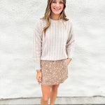 A woman stands against a white textured wall wearing a beige chevron-patterned knit sweater and a brown denim fawn print mini skirt, paired with cream mesh mary jane flats.