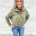 A smiling woman with gray hair stands against a white textured wall wearing an olive green funnel neck cropped utility jacket, blue jeans, and brown boots.