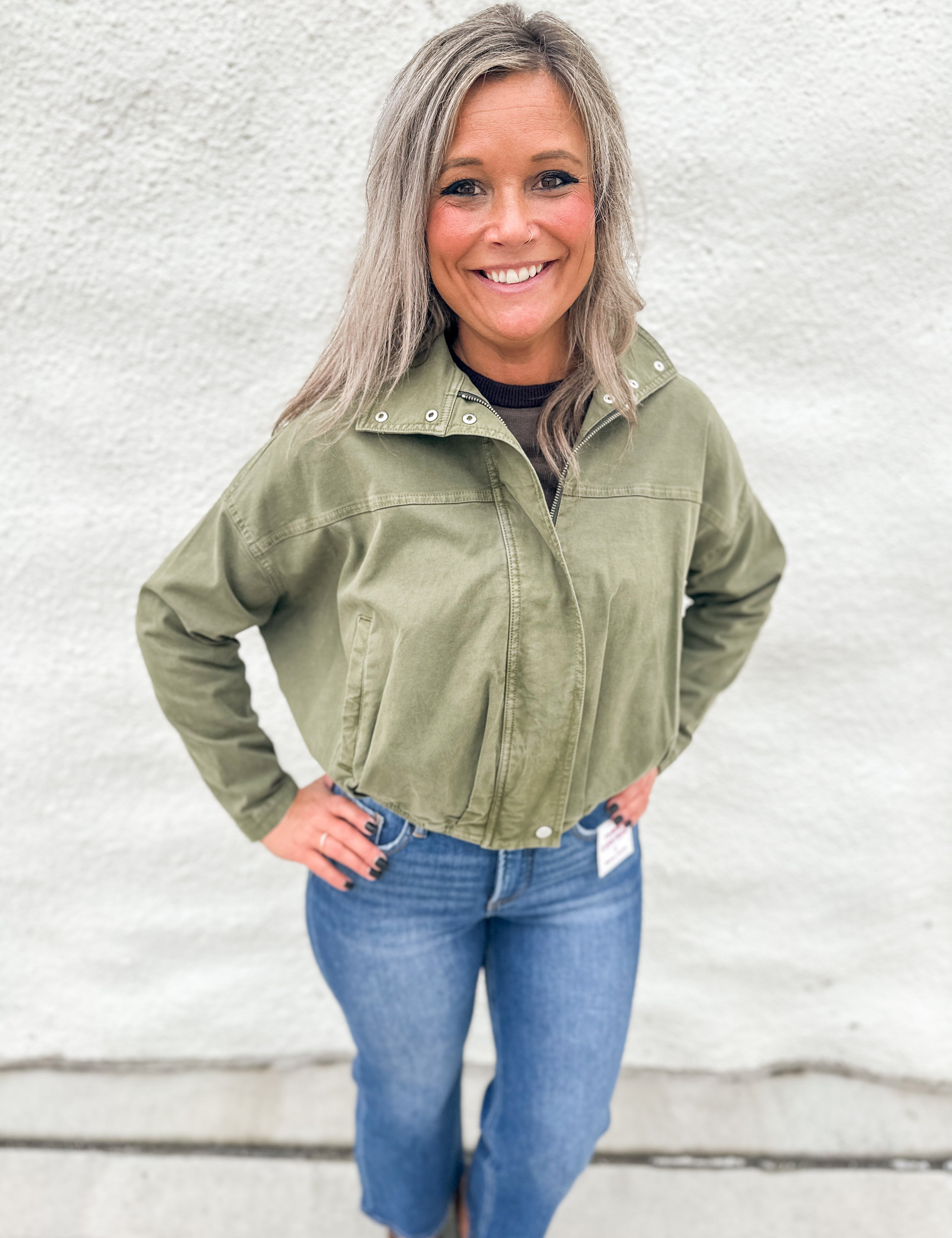 A smiling woman with gray hair stands against a white textured wall wearing an olive green funnel neck cropped utility jacket, blue jeans, and brown boots.