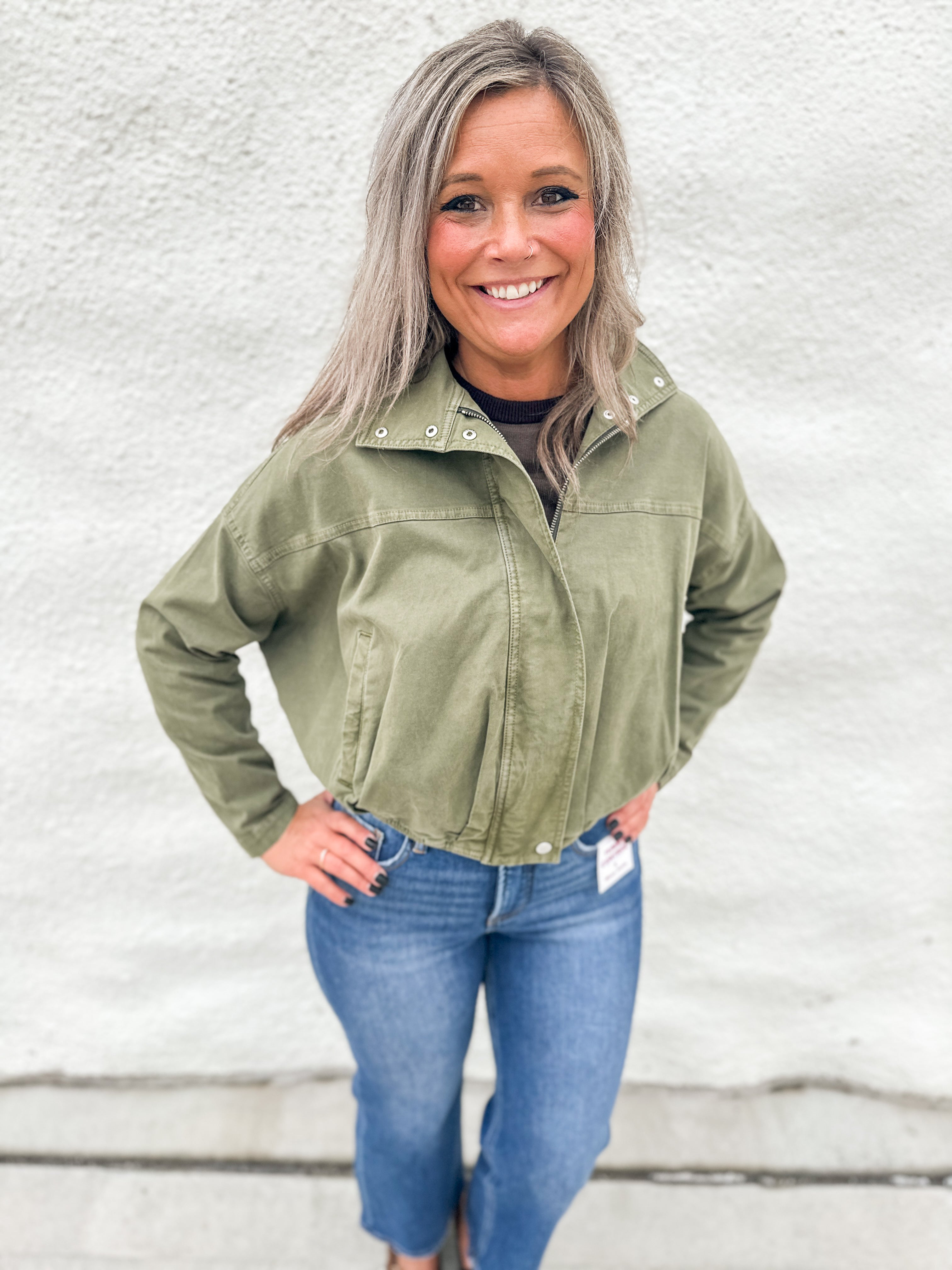 A smiling woman with gray hair stands against a white textured wall wearing an olive green funnel neck cropped utility jacket, blue jeans, and brown boots.