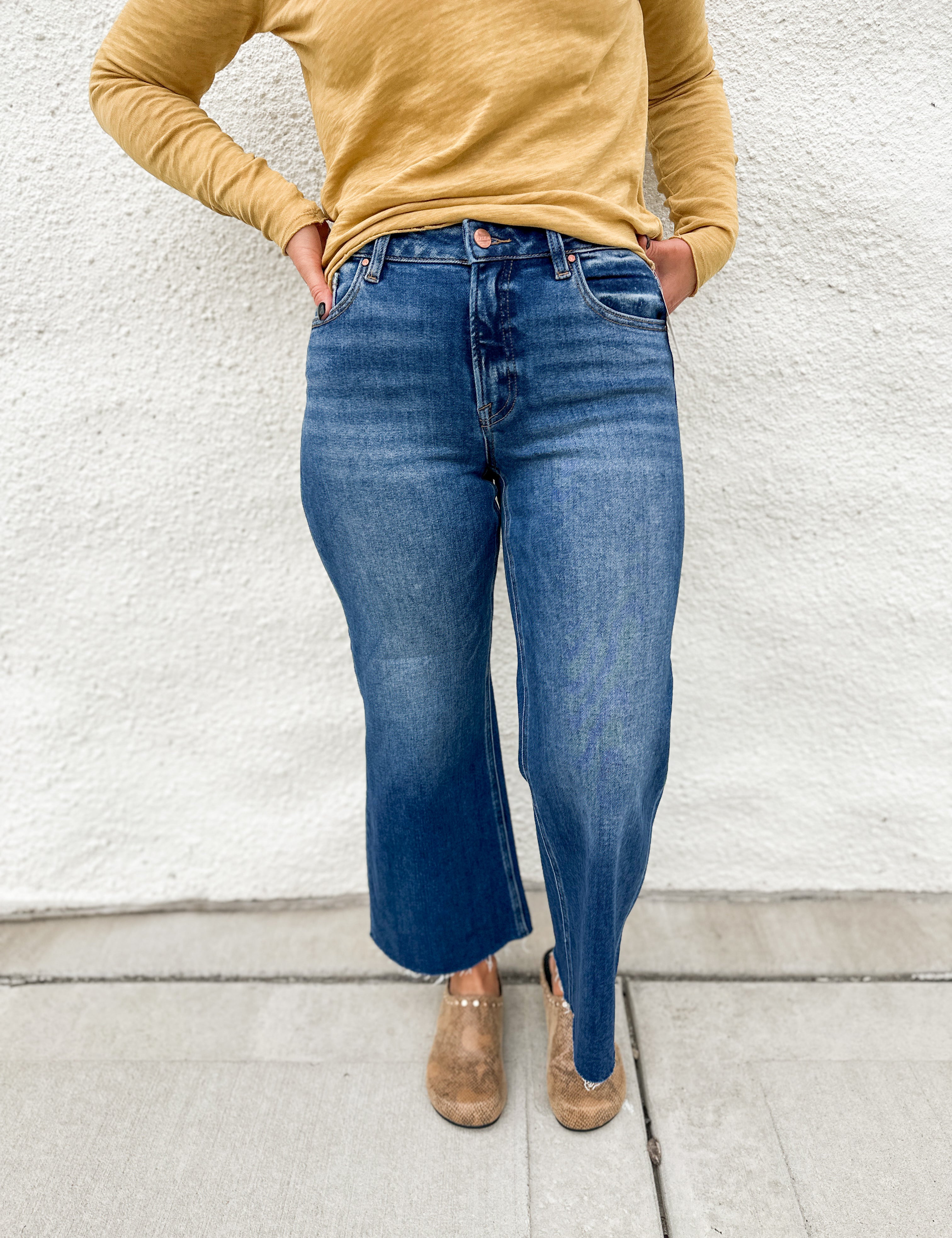 Model wearing a high rise wide leg denim in a dark wash with fading on the legs with a raw hem and gold colored distressed long sleeve tee shirt and faux snakeskin clogs with gold studs