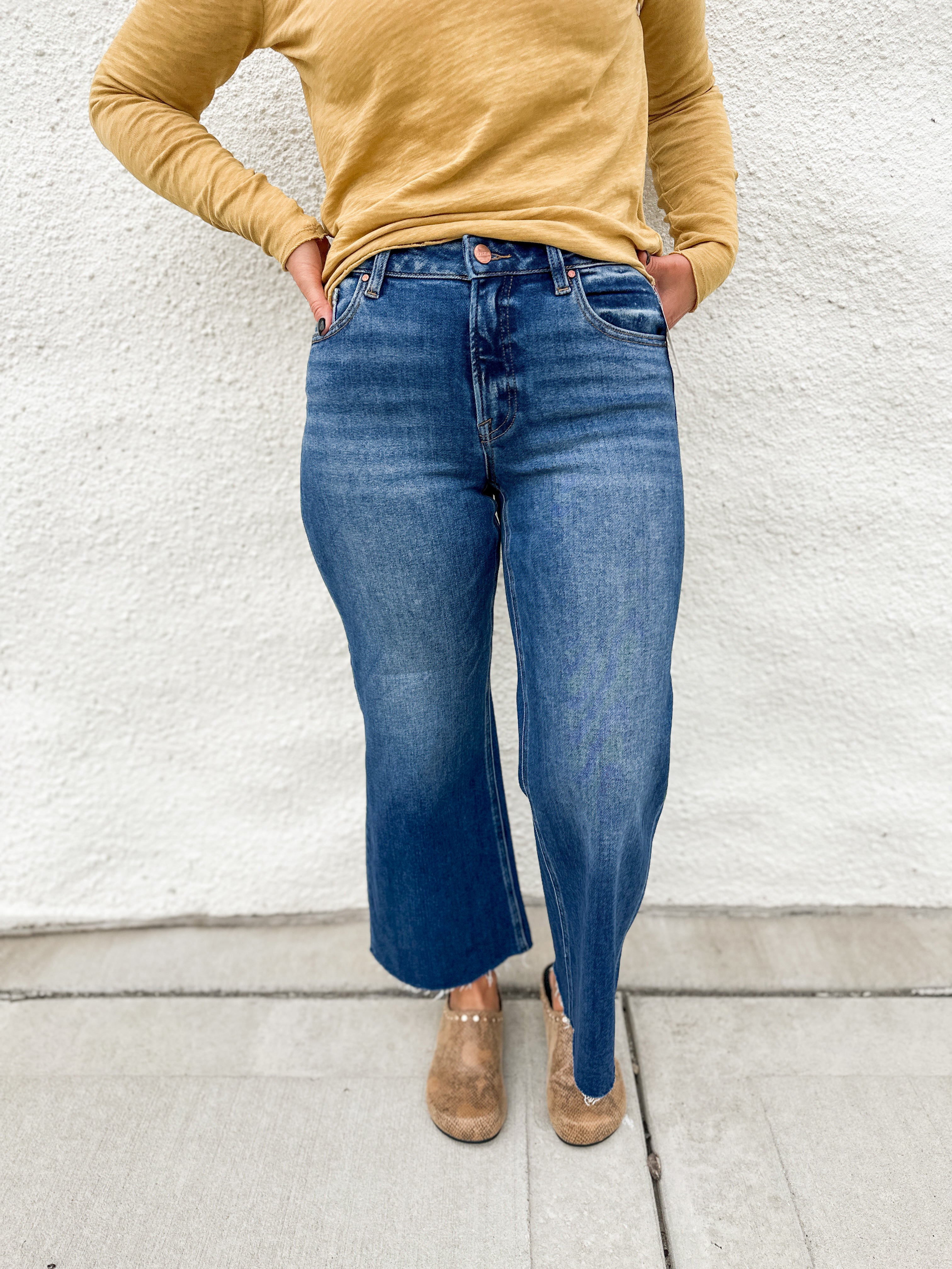 Model wearing a high rise wide leg denim in a dark wash with fading on the legs with a raw hem and gold colored distressed long sleeve tee shirt and faux snakeskin clogs with gold studs