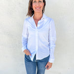 A woman wearing a light blue long-sleeve button-down shirt and blue jeans, standing against a textured off-white wall.