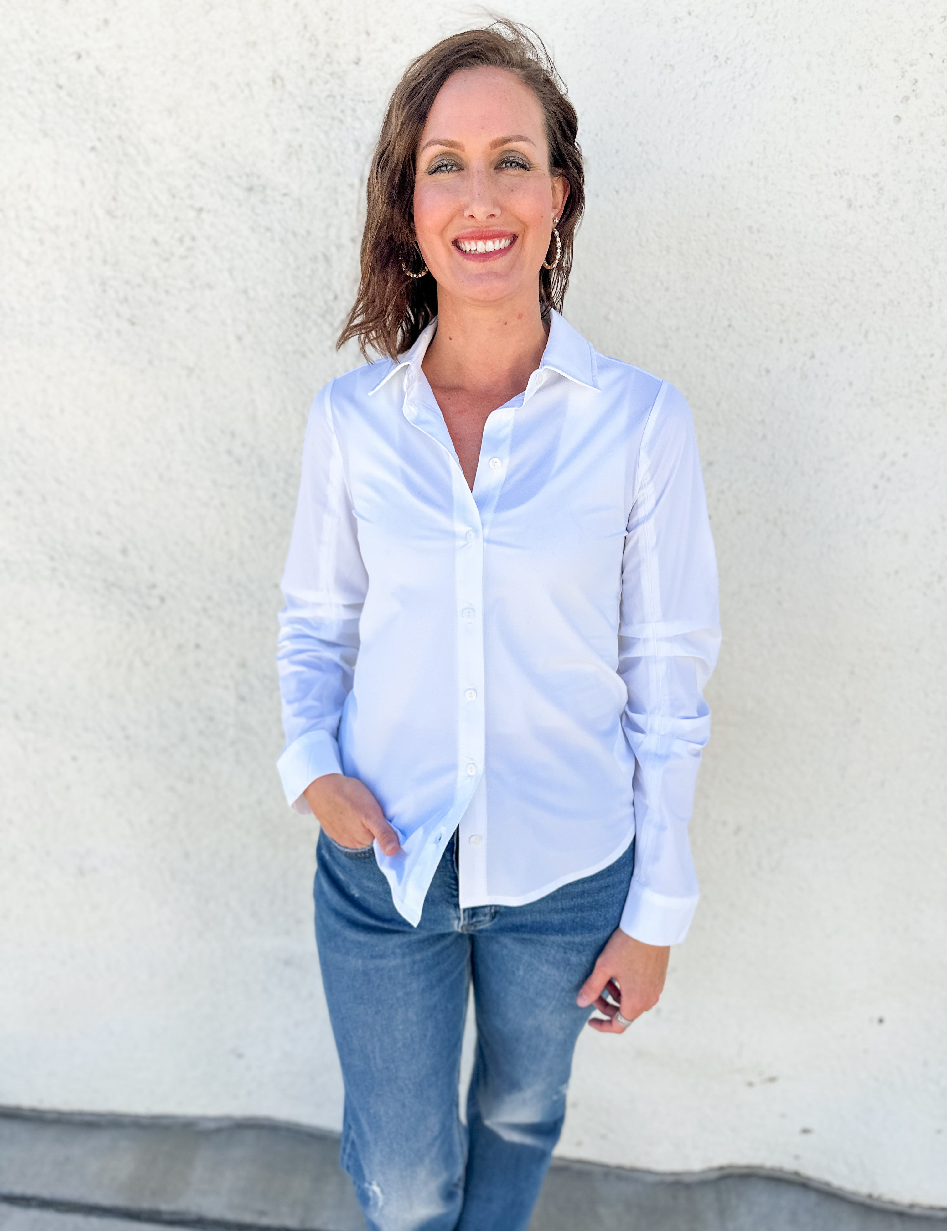 A woman wearing a light blue long-sleeve button-down shirt and blue jeans, standing against a textured off-white wall.