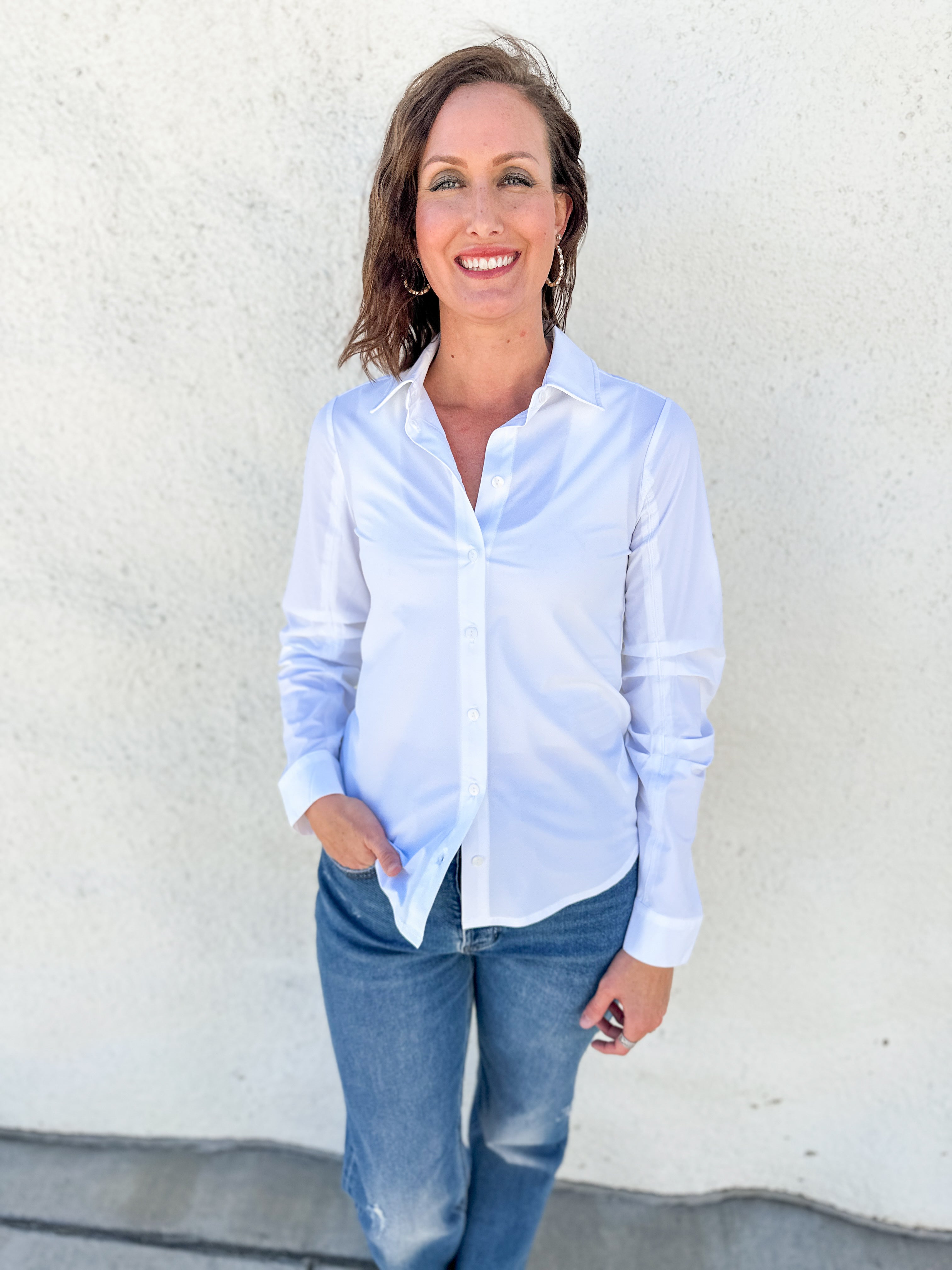 A woman wearing a light blue long-sleeve button-down shirt and blue jeans, standing against a textured off-white wall.
