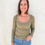 A smiling woman with brown hair stands against a white textured wall wearing an olive green long-sleeve scoop-neck ribbed top and blue jeans.