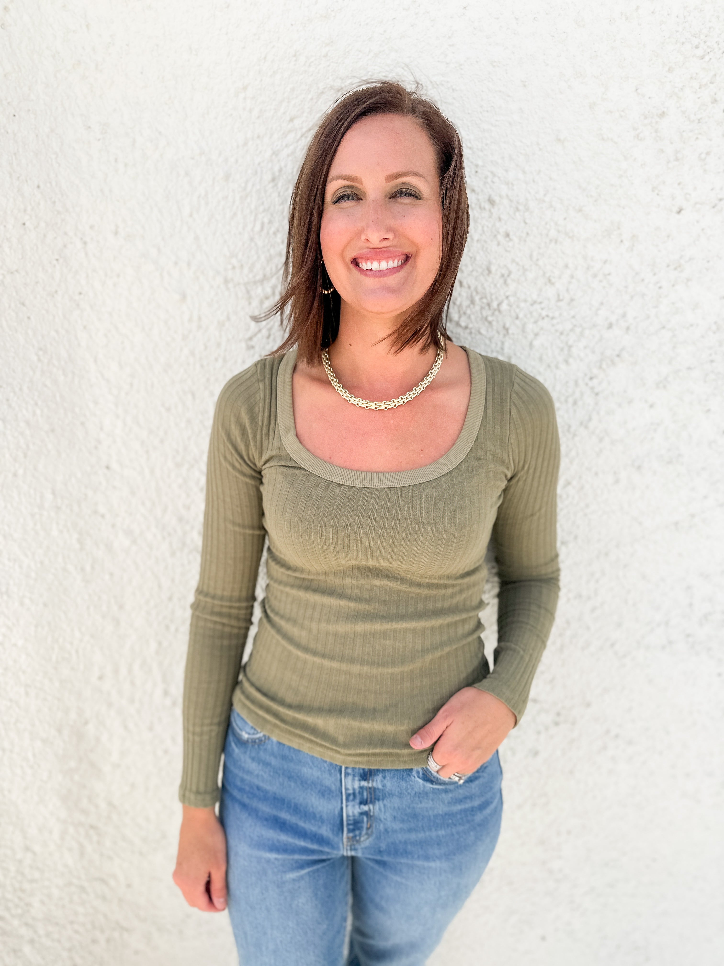 A smiling woman with brown hair stands against a white textured wall wearing an olive green long-sleeve scoop-neck ribbed top and blue jeans.