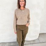 A woman stands outdoors against a white stucco wall wearing a beige ribbed knit sweater by Dear John Denim, olive green pants, and tan driving moccasins with stud details.