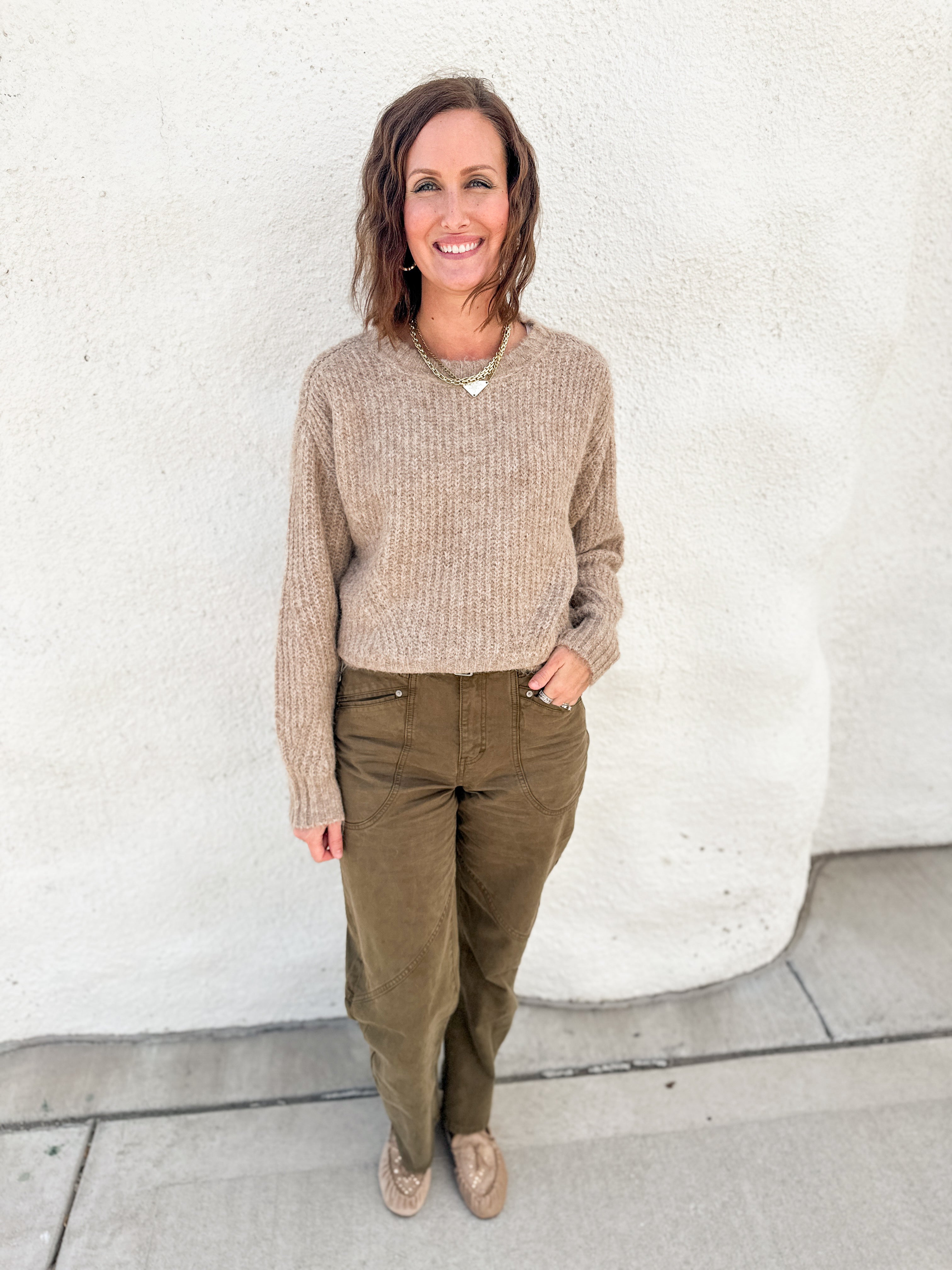 A woman stands outdoors against a white stucco wall wearing a beige ribbed knit sweater by Dear John Denim, olive green pants, and tan driving moccasins with stud details.