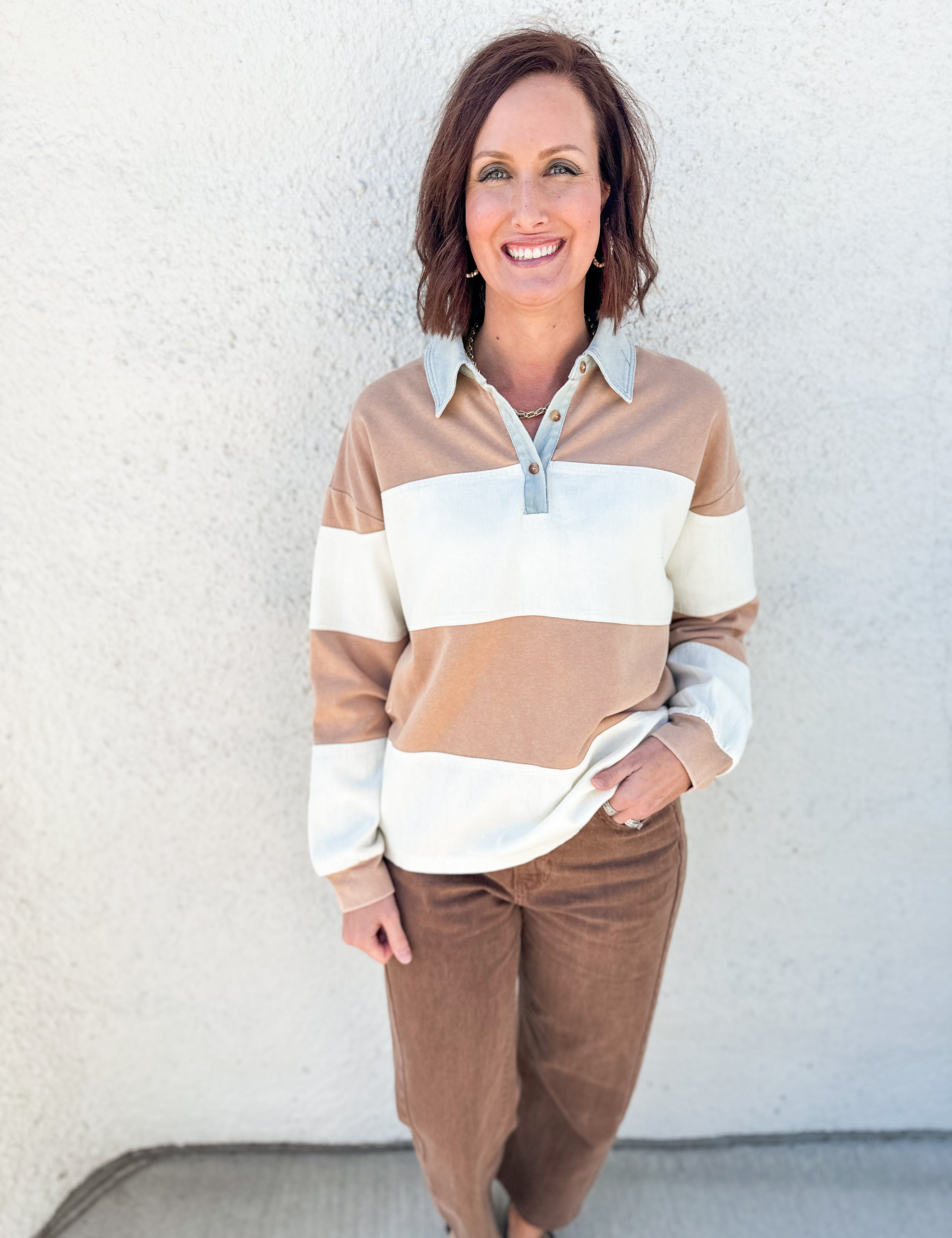 A woman wearing a beige, white, and brown striped long-sleeve polo shirt with brown barrel leg pants, standing against a textured white stucco wall.