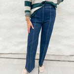 A person wearing dark blue straight-leg jeans with front seaming and elastic waistband from Liverpool, a green and cream striped long-sleeve shirt from Thread & Supply, and cream mesh Mary Jane slip-on shoes, standing against a white textured wall