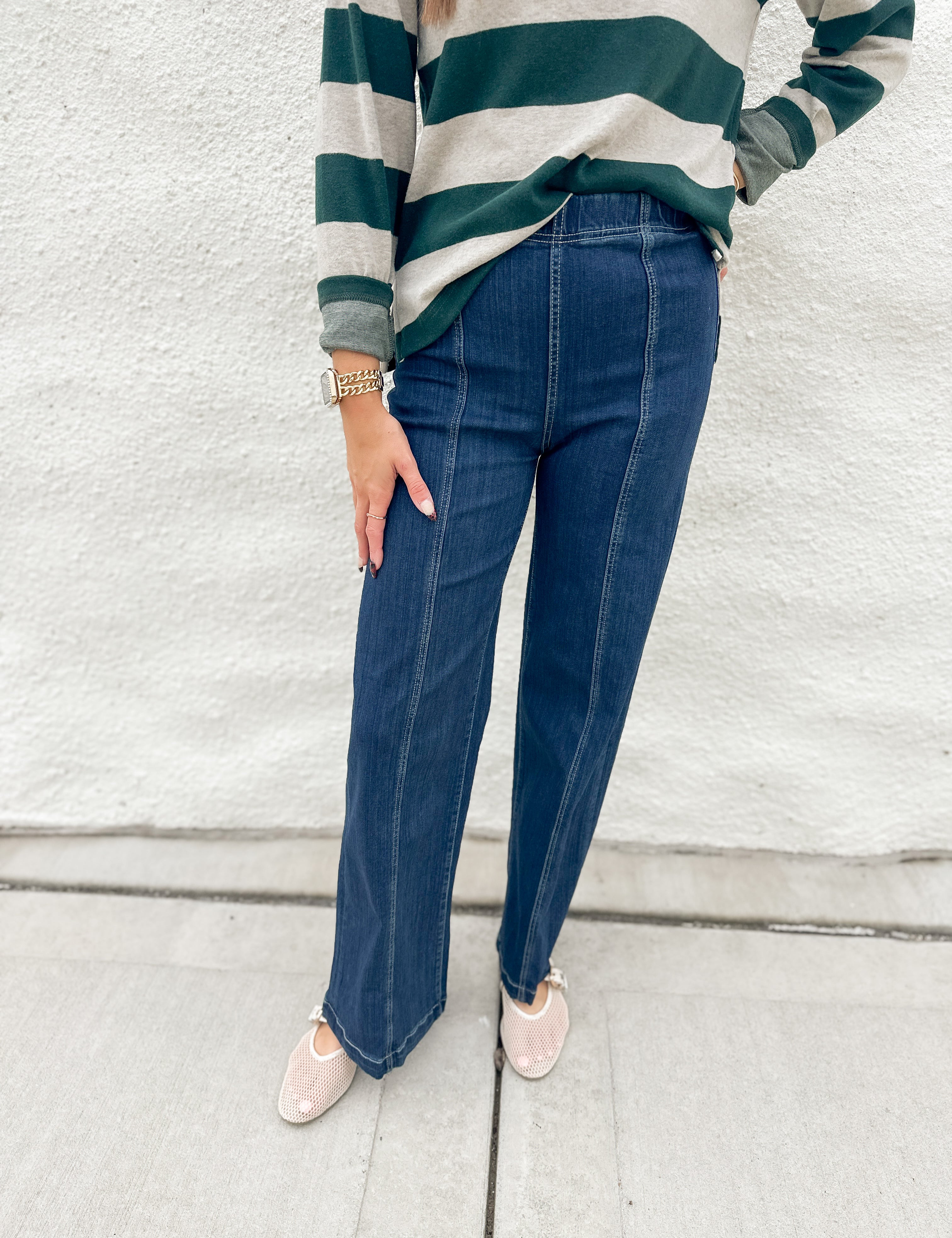 A person wearing dark blue straight-leg jeans with front seaming and elastic waistband from Liverpool, a green and cream striped long-sleeve shirt from Thread & Supply, and cream mesh Mary Jane slip-on shoes, standing against a white textured wall