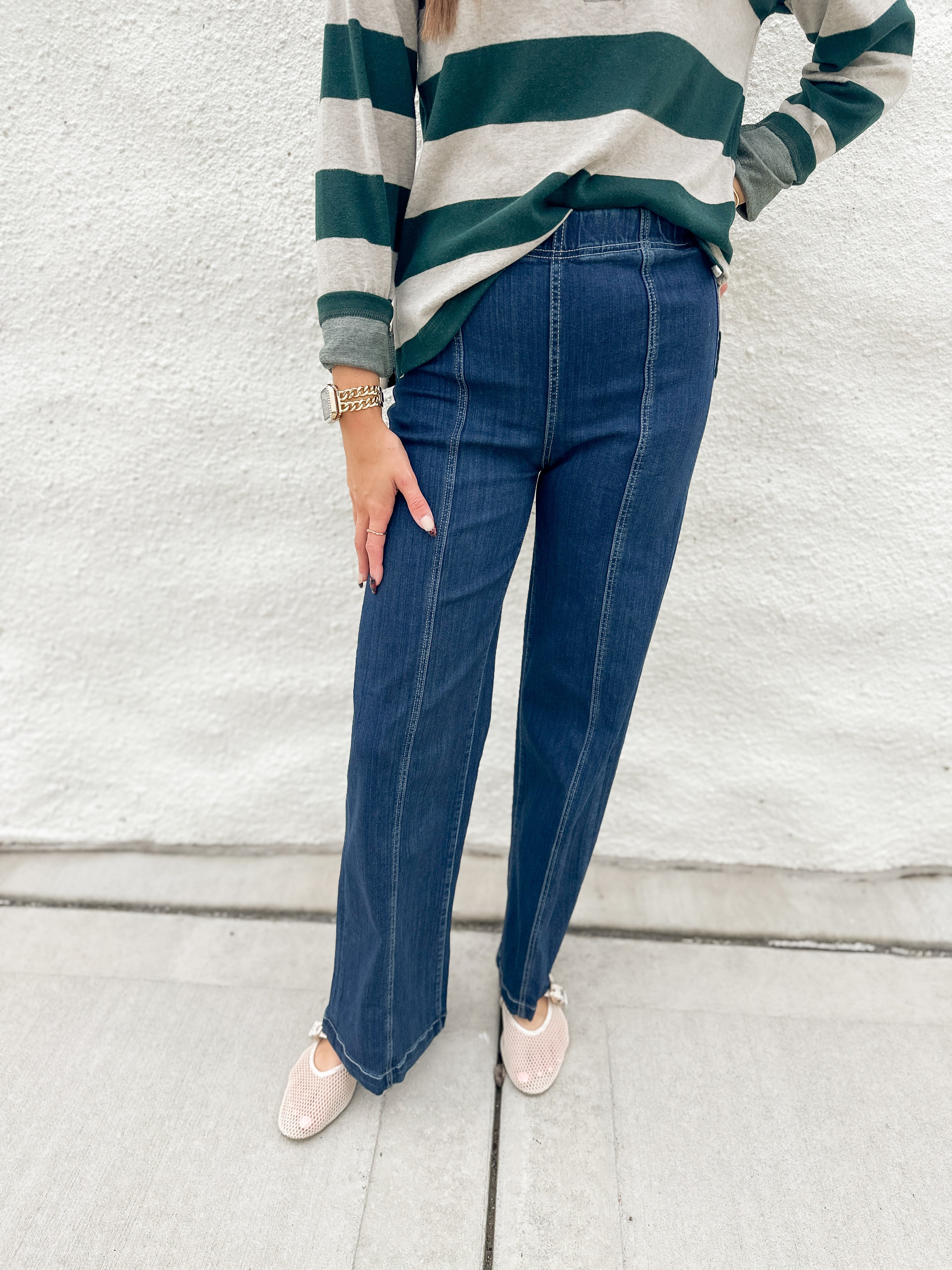 A person wearing dark blue straight-leg jeans with front seaming and elastic waistband from Liverpool, a green and cream striped long-sleeve shirt from Thread & Supply, and cream mesh Mary Jane slip-on shoes, standing against a white textured wall