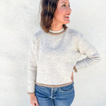 A woman wearing a light oatmeal gray melange heathered knit sweater with beige ribbed trim, paired with blue jeans, standing against a white stucco wall.