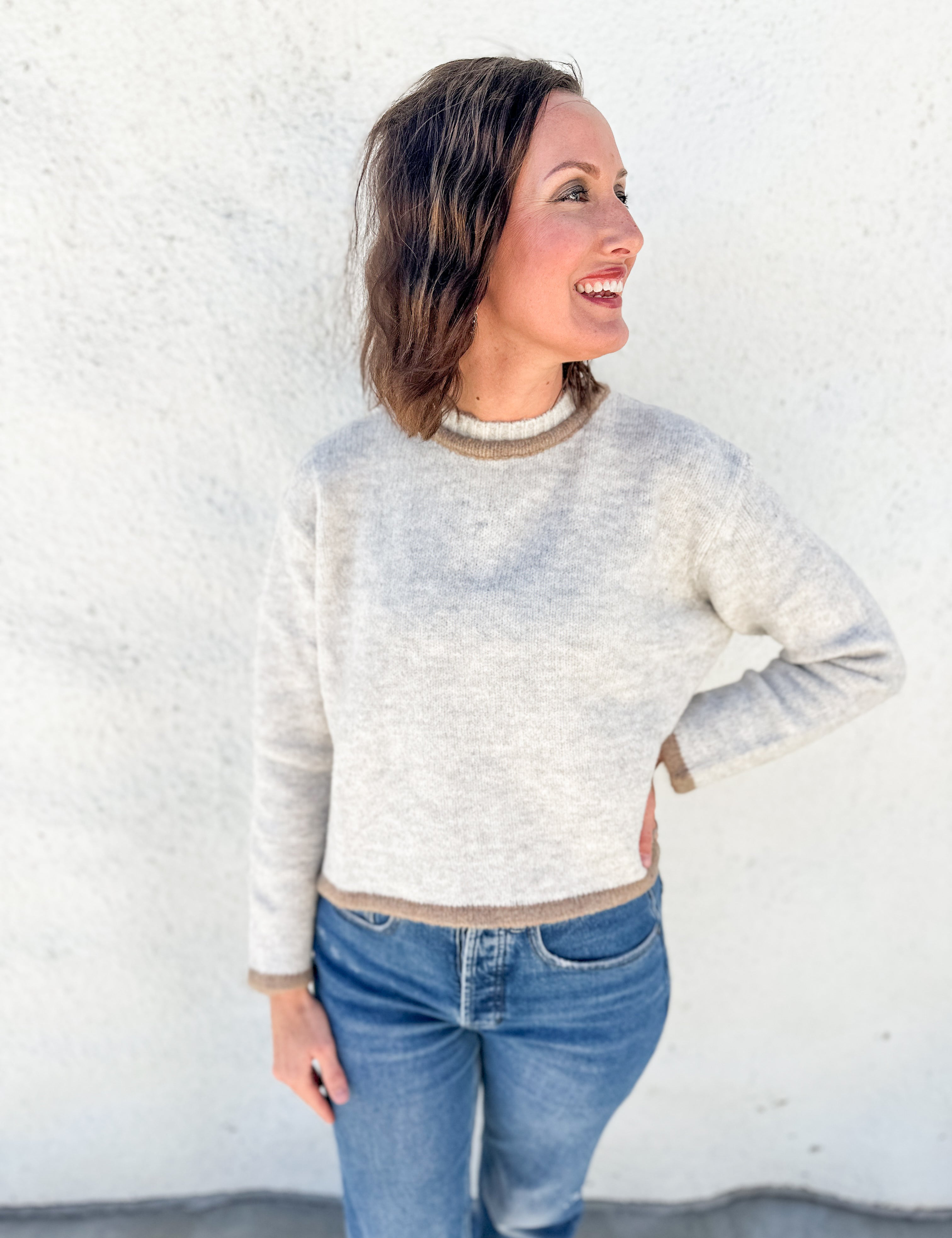A woman wearing a light oatmeal gray melange heathered knit sweater with beige ribbed trim, paired with blue jeans, standing against a white stucco wall.
