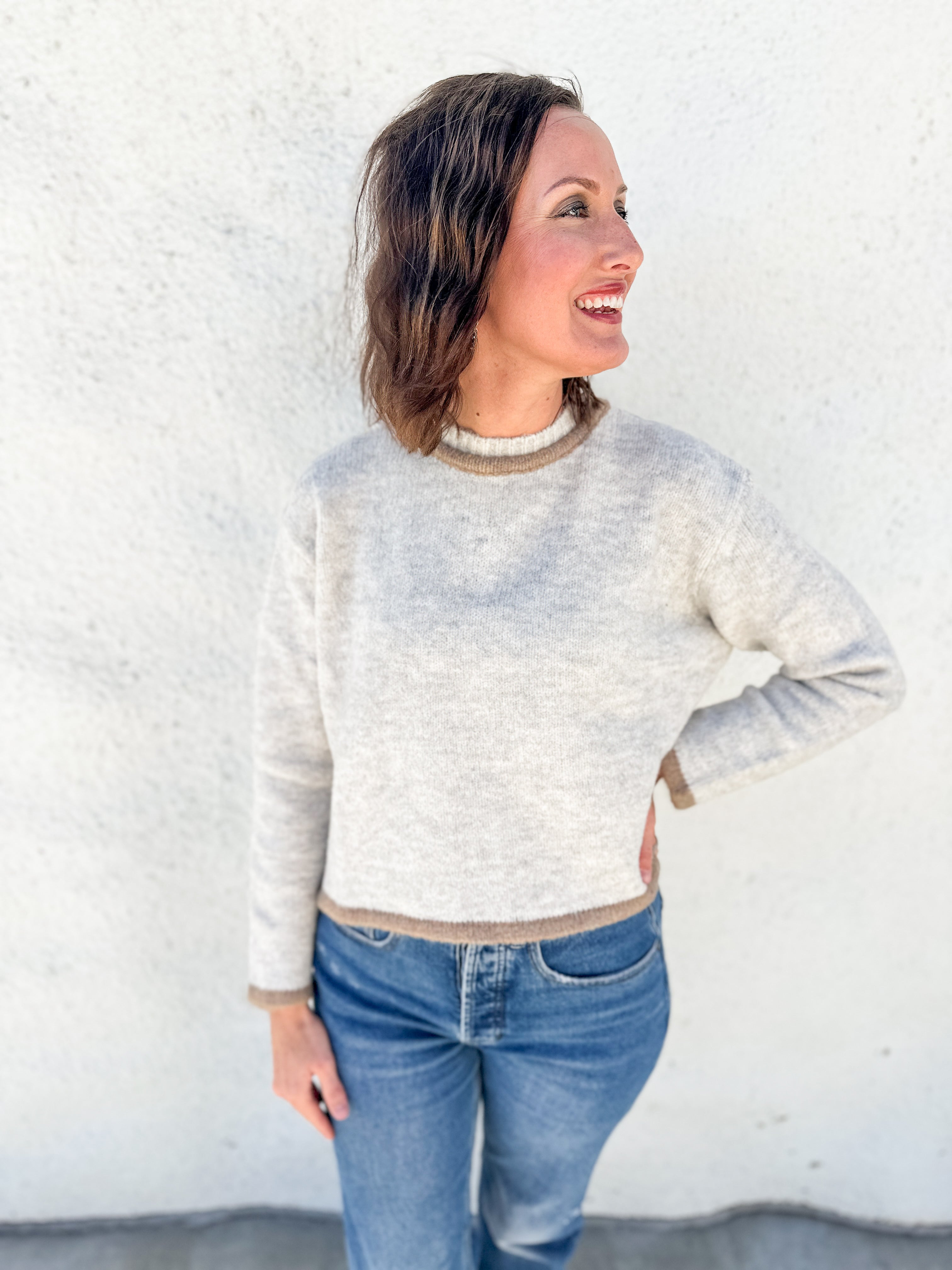 A woman wearing a light oatmeal gray melange heathered knit sweater with beige ribbed trim, paired with blue jeans, standing against a white stucco wall.