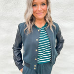 A woman wearing a black and green bomber jacket with faux leather sleeves over a green and white striped textured tee and wearing green wide leg jogger pants against a white wall