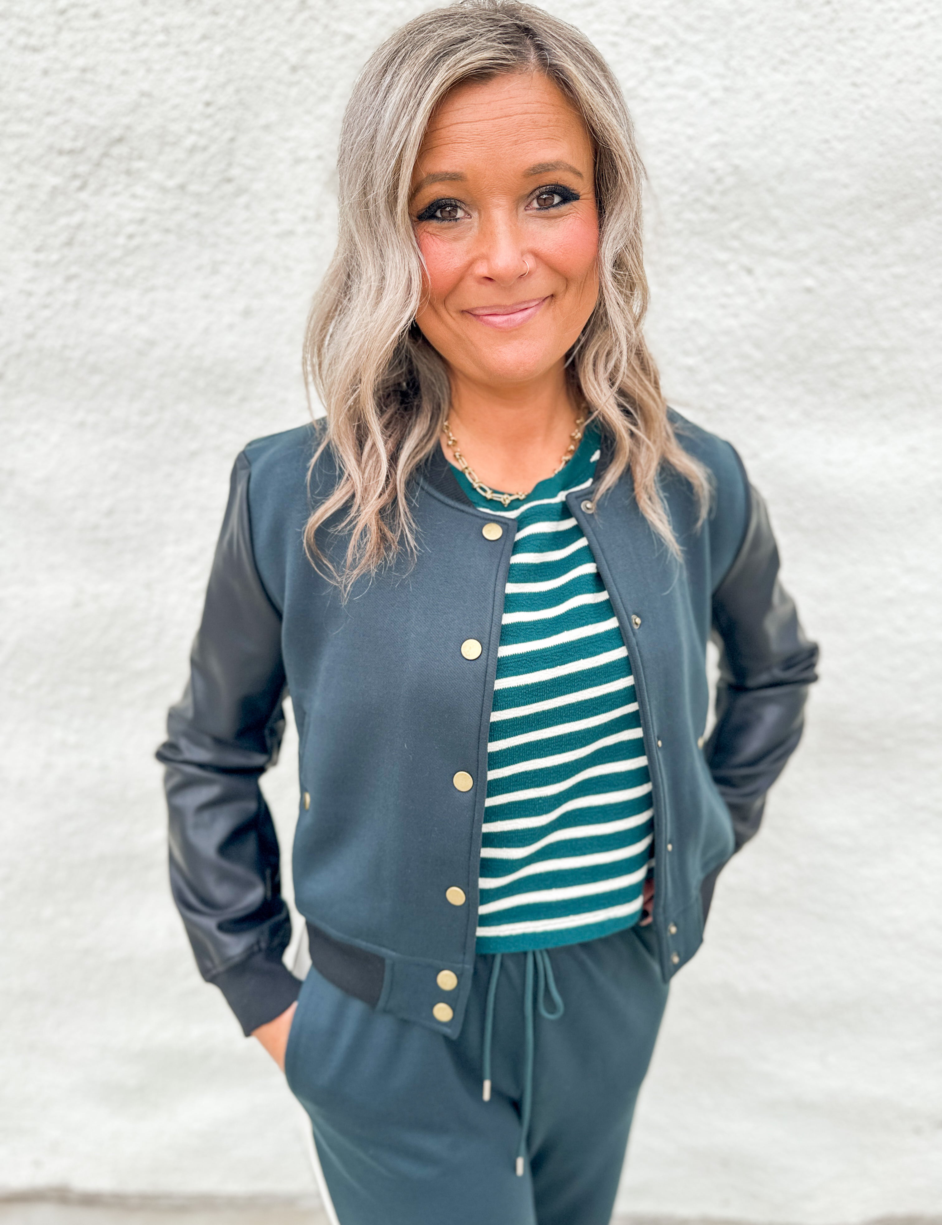 A woman wearing a black and green bomber jacket with faux leather sleeves over a green and white striped textured tee and wearing green wide leg jogger pants against a white wall