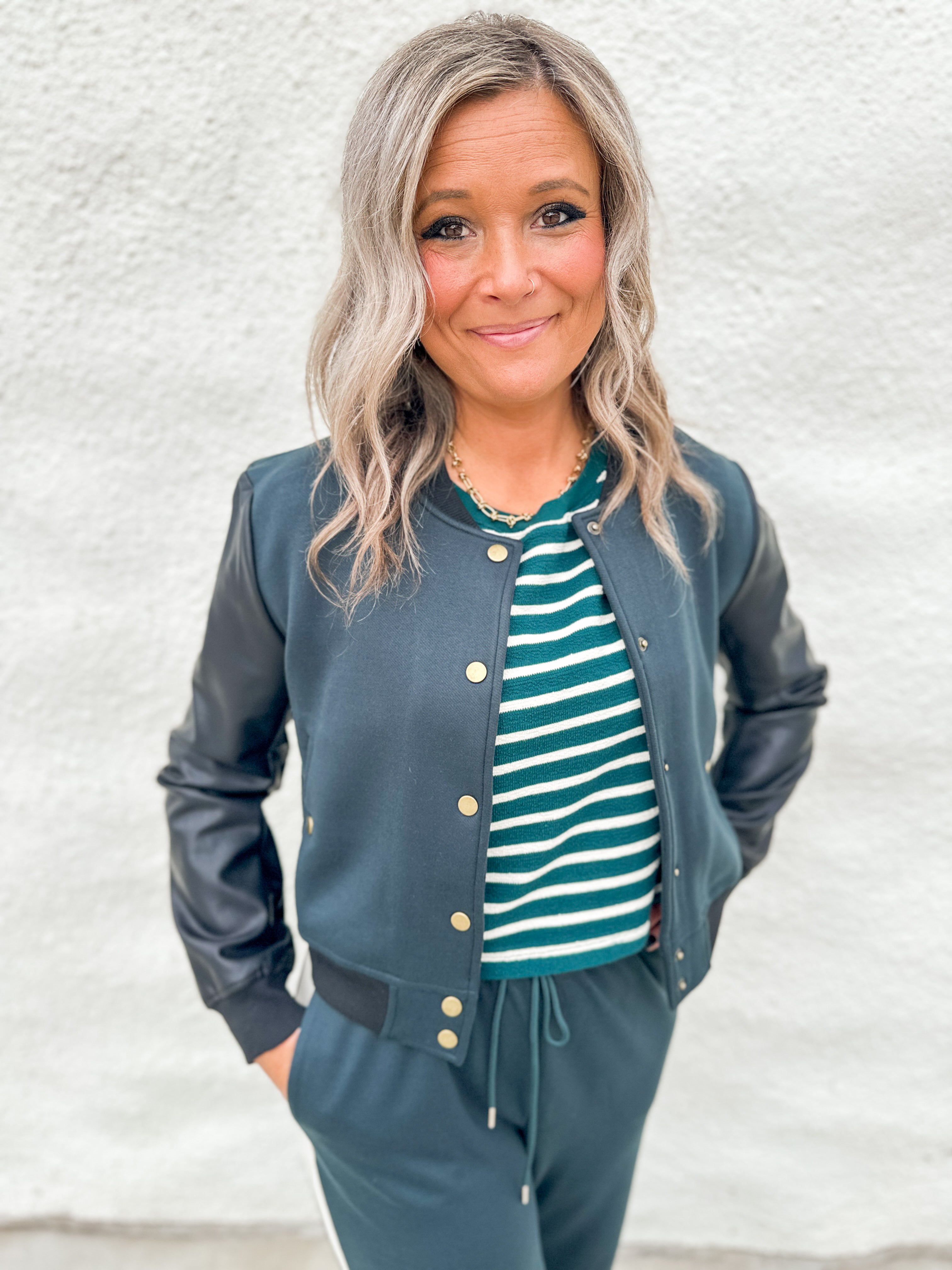 A woman wearing a black and green bomber jacket with faux leather sleeves over a green and white striped textured tee and wearing green wide leg jogger pants against a white wall