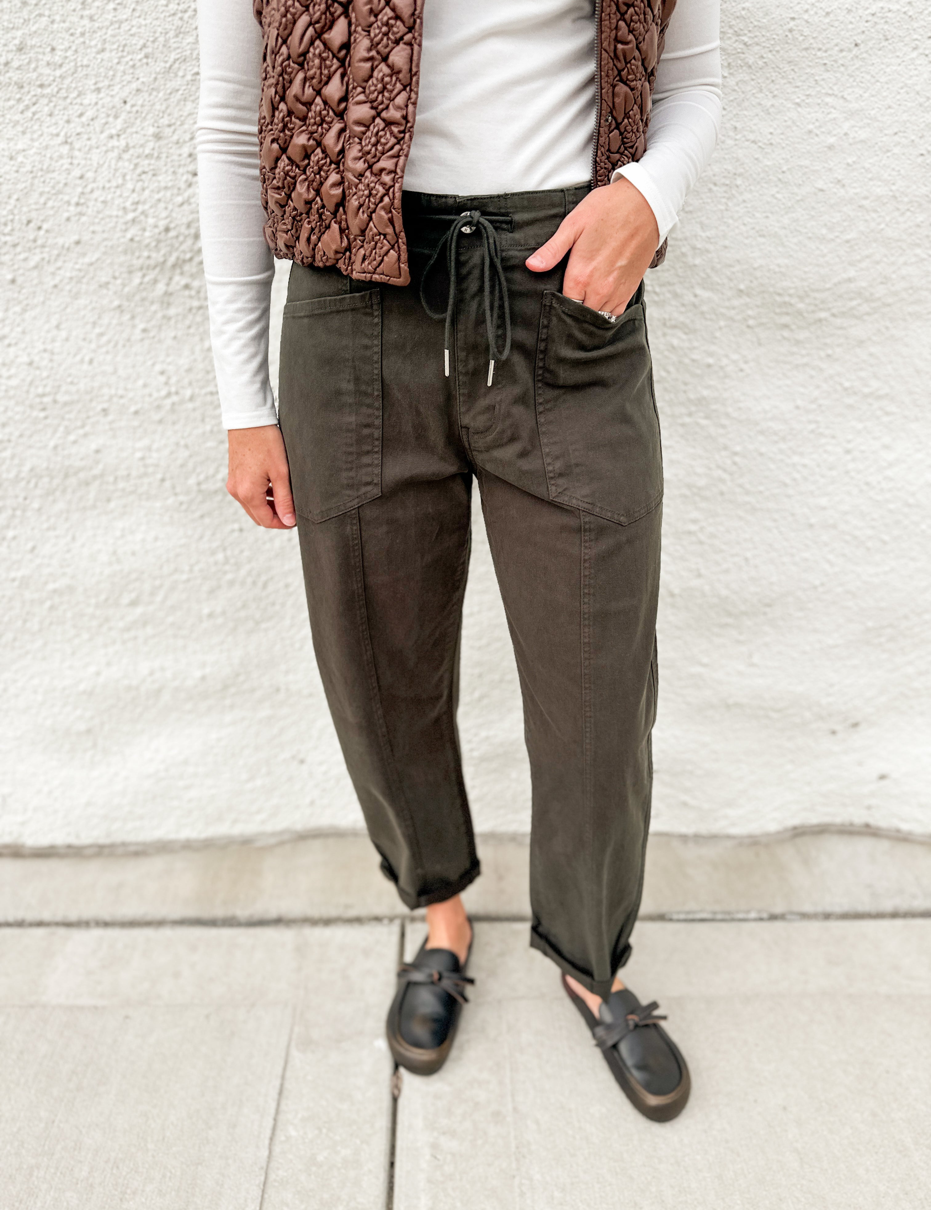 Dark green barrel denim with front pockets and drawstring waistband, long sleeve lightly ribbed long sleeve top and brown faux leather look vest with stitched details, zippered front and buttons, distressed black leather clogs