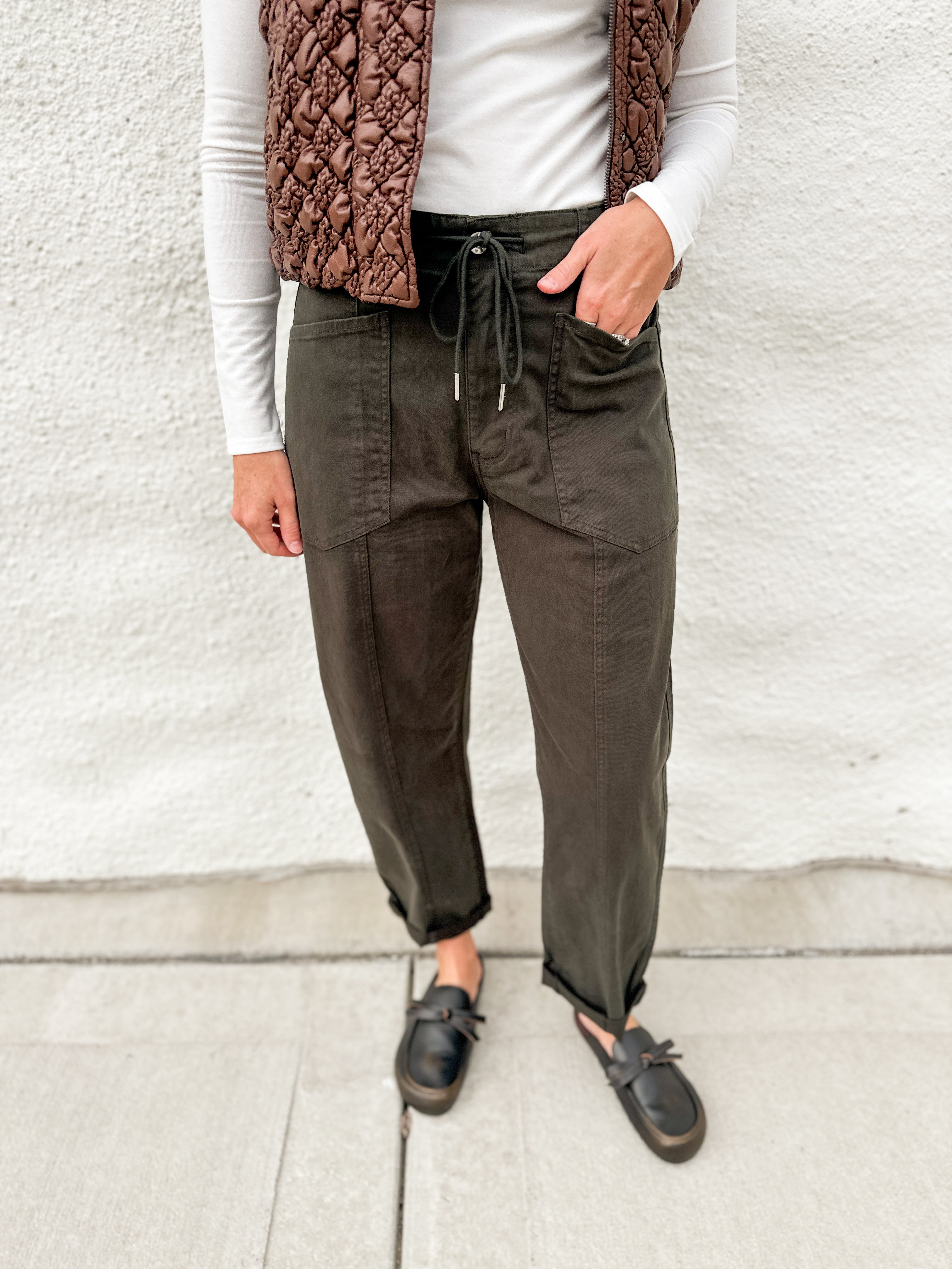 Dark green barrel denim with front pockets and drawstring waistband, long sleeve lightly ribbed long sleeve top and brown faux leather look vest with stitched details, zippered front and buttons, distressed black leather clogs
