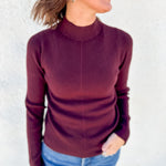 A woman wearing a ribbed mock neck sweater in burgundy, paired with blue jeans, standing against a light-colored textured wall.