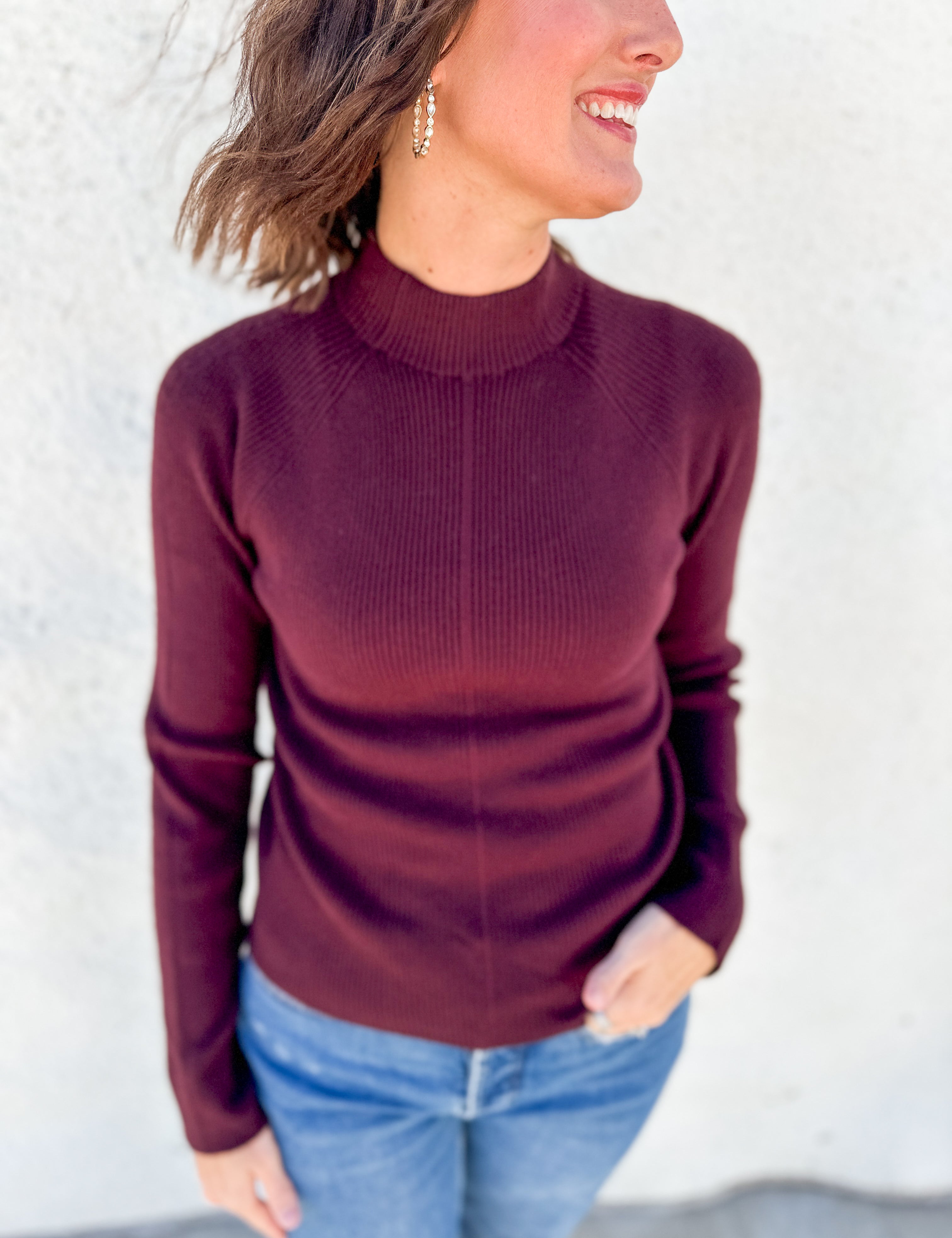 A woman wearing a ribbed mock neck sweater in burgundy, paired with blue jeans, standing against a light-colored textured wall.