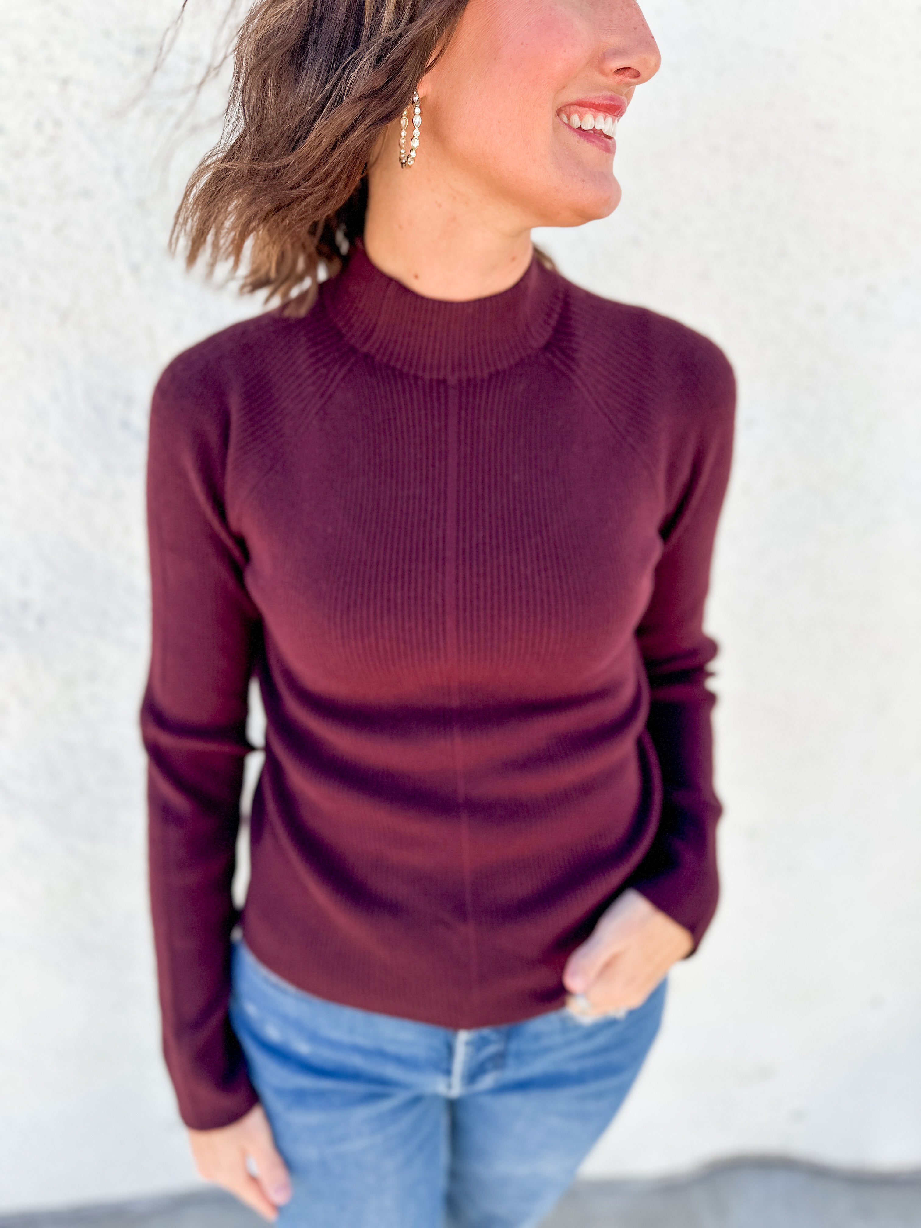 A woman wearing a ribbed mock neck sweater in burgundy, paired with blue jeans, standing against a light-colored textured wall.