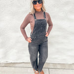 A woman wearing dark gray denim overall over a taupe long-sleeve top, styled with black sunglasses and brown leather sandals, standing against a white stucco wall.