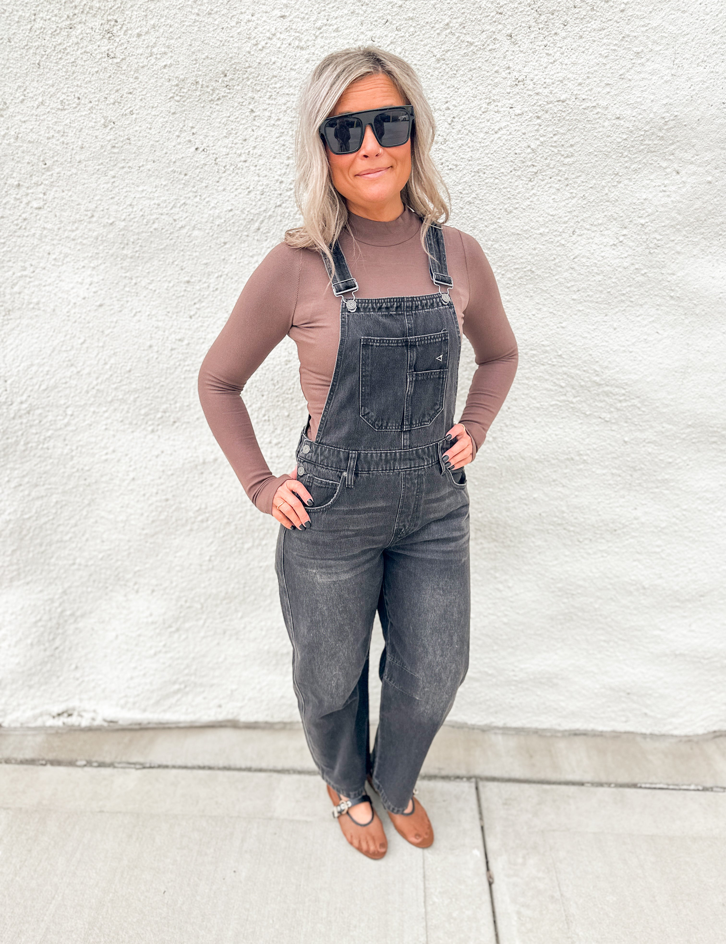 A woman wearing dark gray denim overall over a taupe long-sleeve top, styled with black sunglasses and brown leather sandals, standing against a white stucco wall.