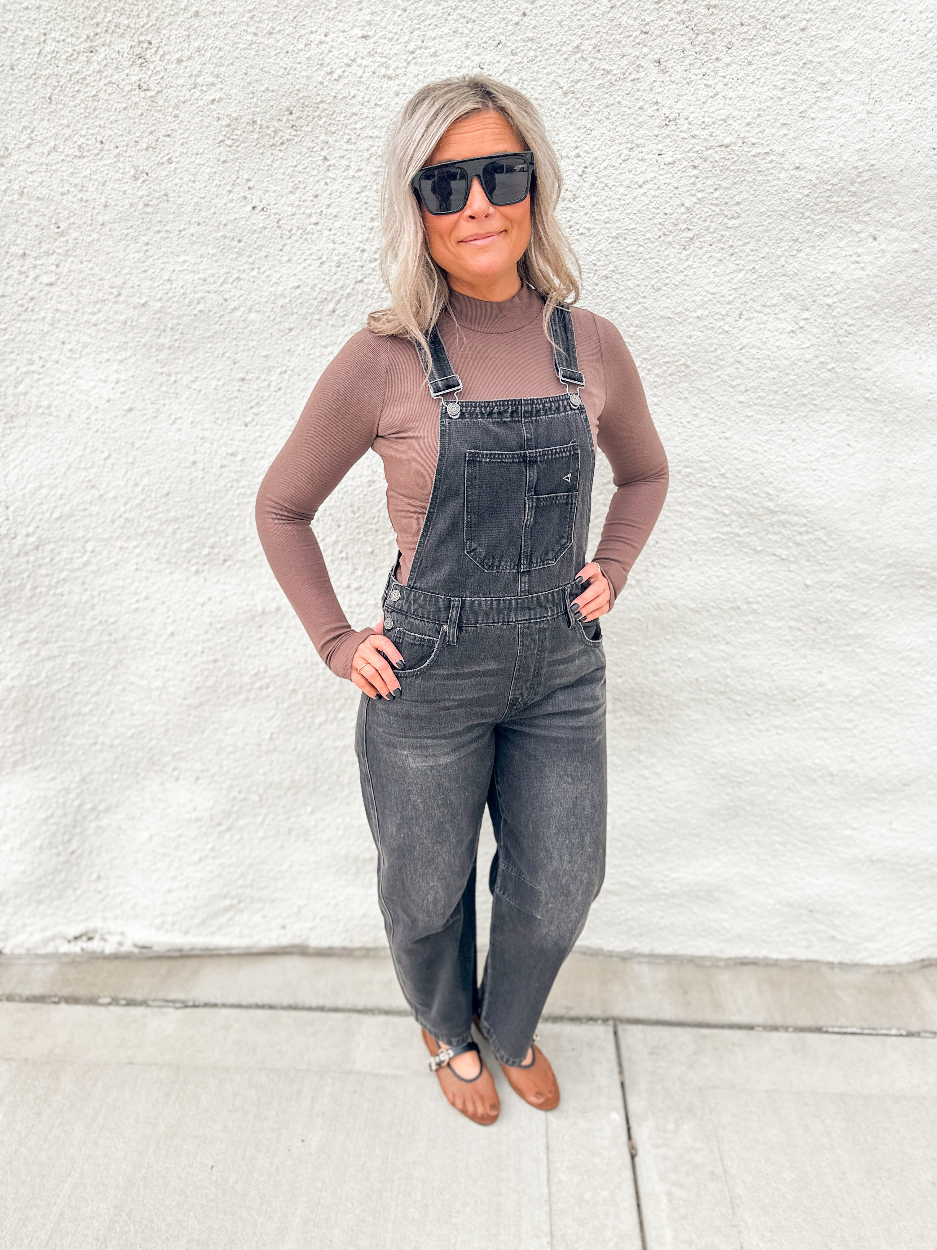 A woman wearing dark gray denim overall over a taupe long-sleeve top, styled with black sunglasses and brown leather sandals, standing against a white stucco wall.