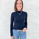 A woman with shoulder-length brown hair stands against a textured white wall, wearing a fitted black long-sleeve mock neck top and light blue jeans.
