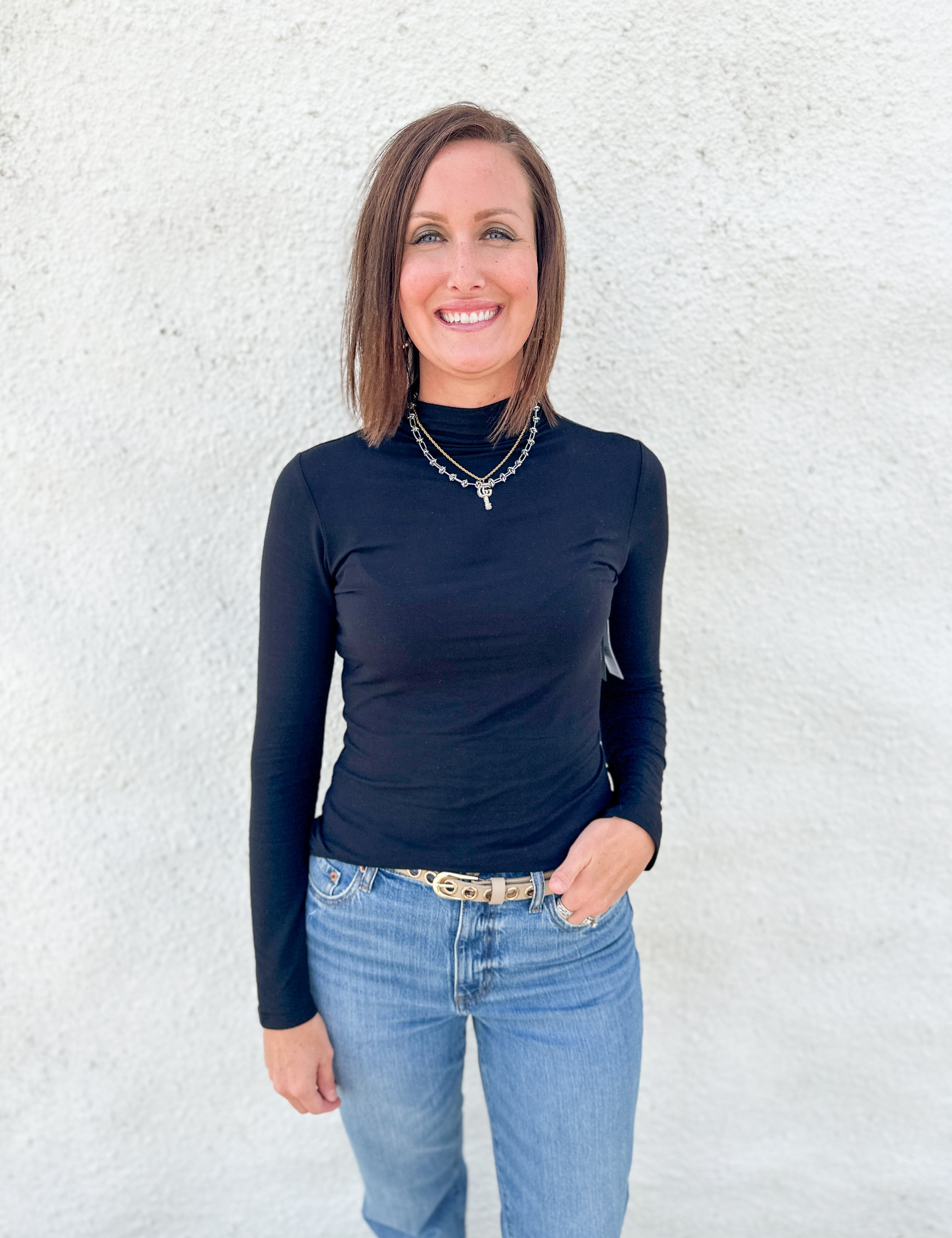 A woman with shoulder-length brown hair stands against a textured white wall, wearing a fitted black long-sleeve mock neck top and light blue jeans.