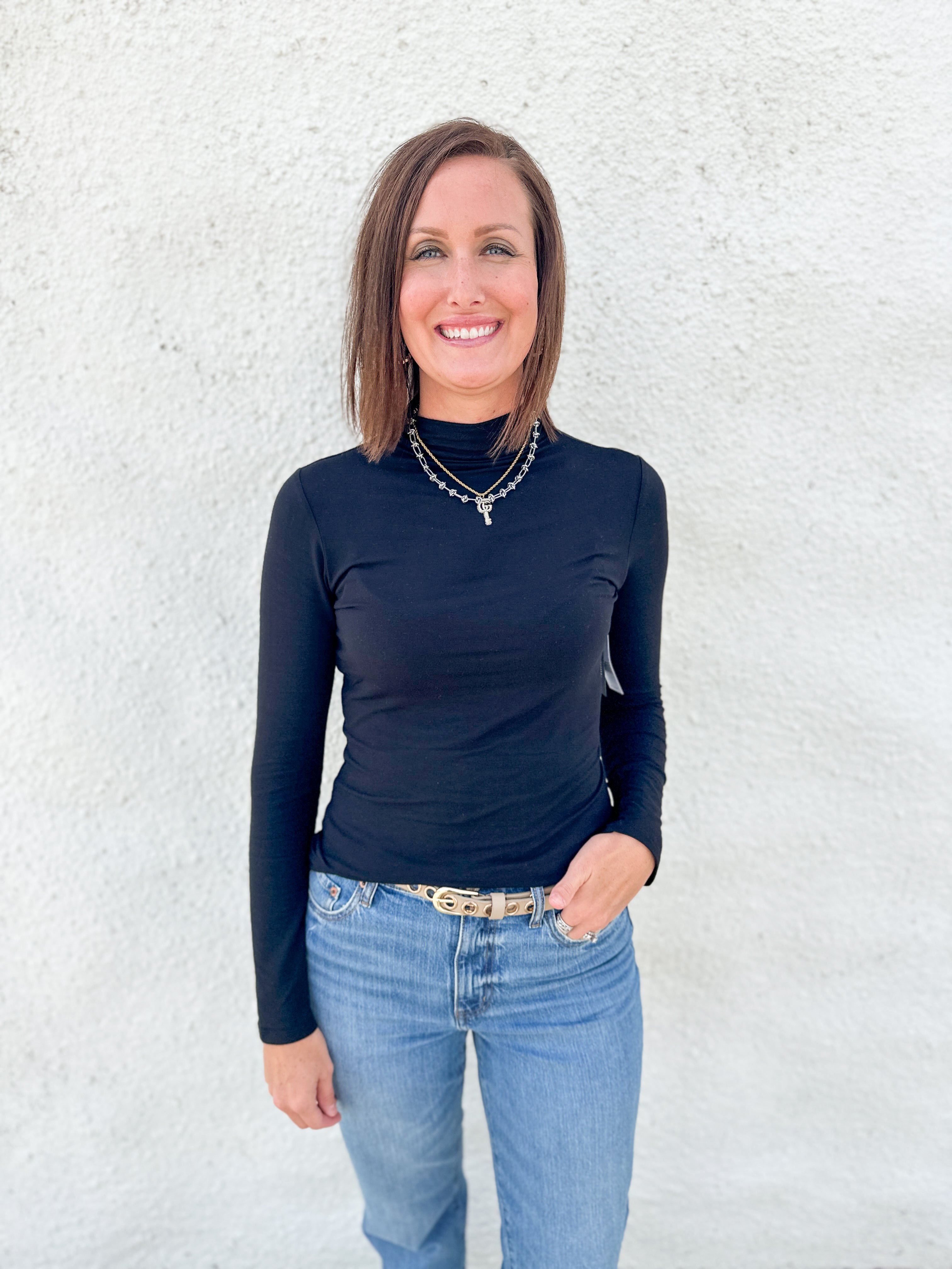 A woman with shoulder-length brown hair stands against a textured white wall, wearing a fitted black long-sleeve mock neck top and light blue jeans.