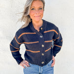 A woman wearing a navy blue chunky knit cardigan with brown stripes and brown buttons, paired with light blue jeans, standing against a white stucco wall.