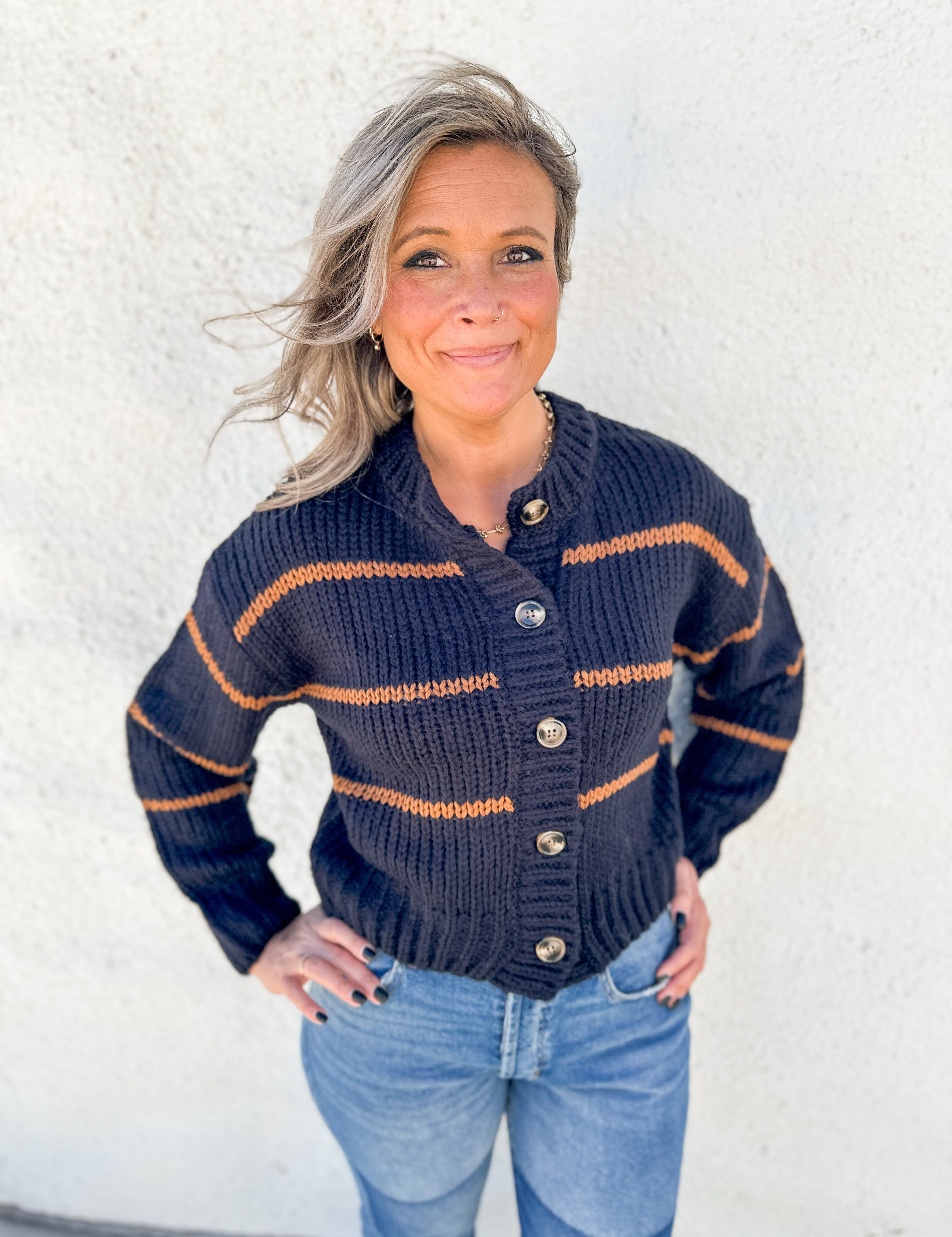 A woman wearing a navy blue chunky knit cardigan with brown stripes and brown buttons, paired with light blue jeans, standing against a white stucco wall.