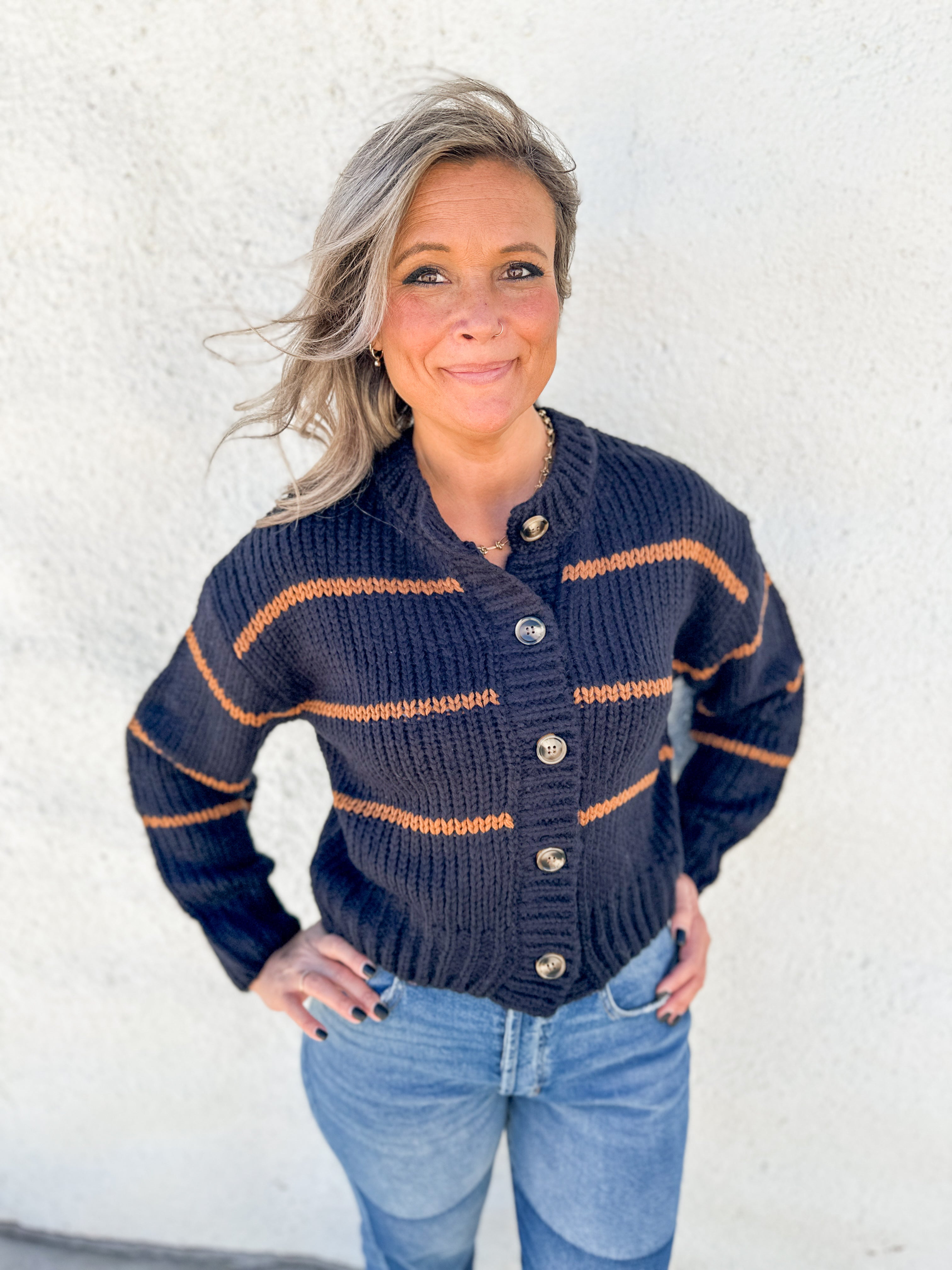 A woman wearing a navy blue chunky knit cardigan with brown stripes and brown buttons, paired with light blue jeans, standing against a white stucco wall.