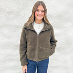 A woman smiling while wearing an olive brown fleece zip-up jacket from Kut from the Kloth over a white henley shirt and blue jeans, standing against a white textured wall.